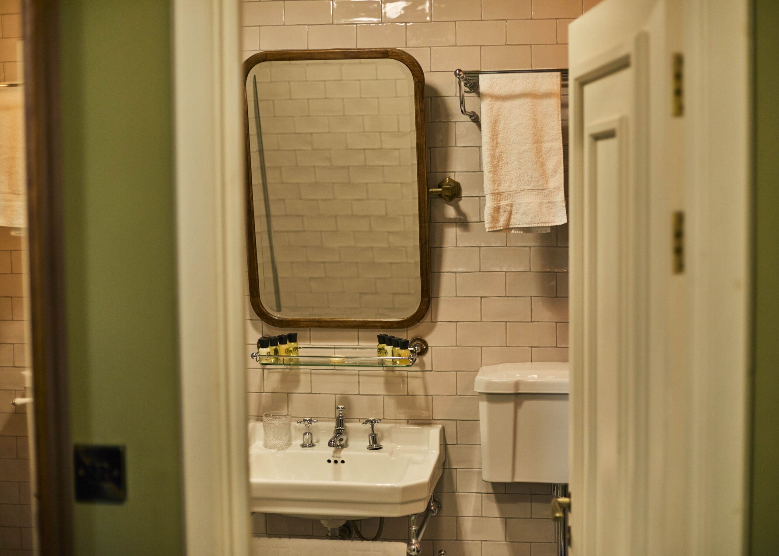 A bathroom with a white sink, a rectangular mirror above it, a glass shelf with toiletries, a towel hanging on a rack, and a toilet partially visible behind an open door. The walls are tiled.