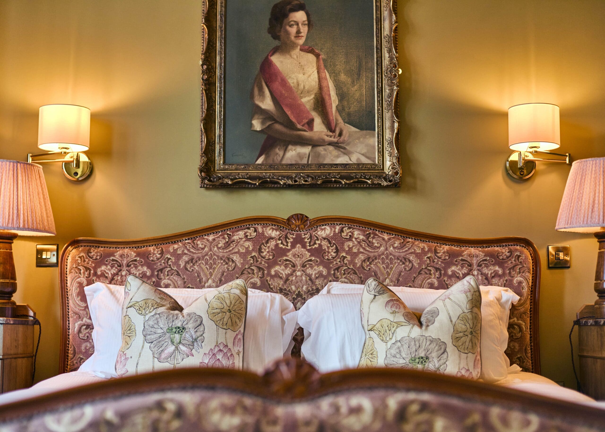 A vintage bedroom features an ornate pink upholstered headboard, floral pillows, bedside lamps, and a framed portrait of a woman in formal attire hanging above the bed.