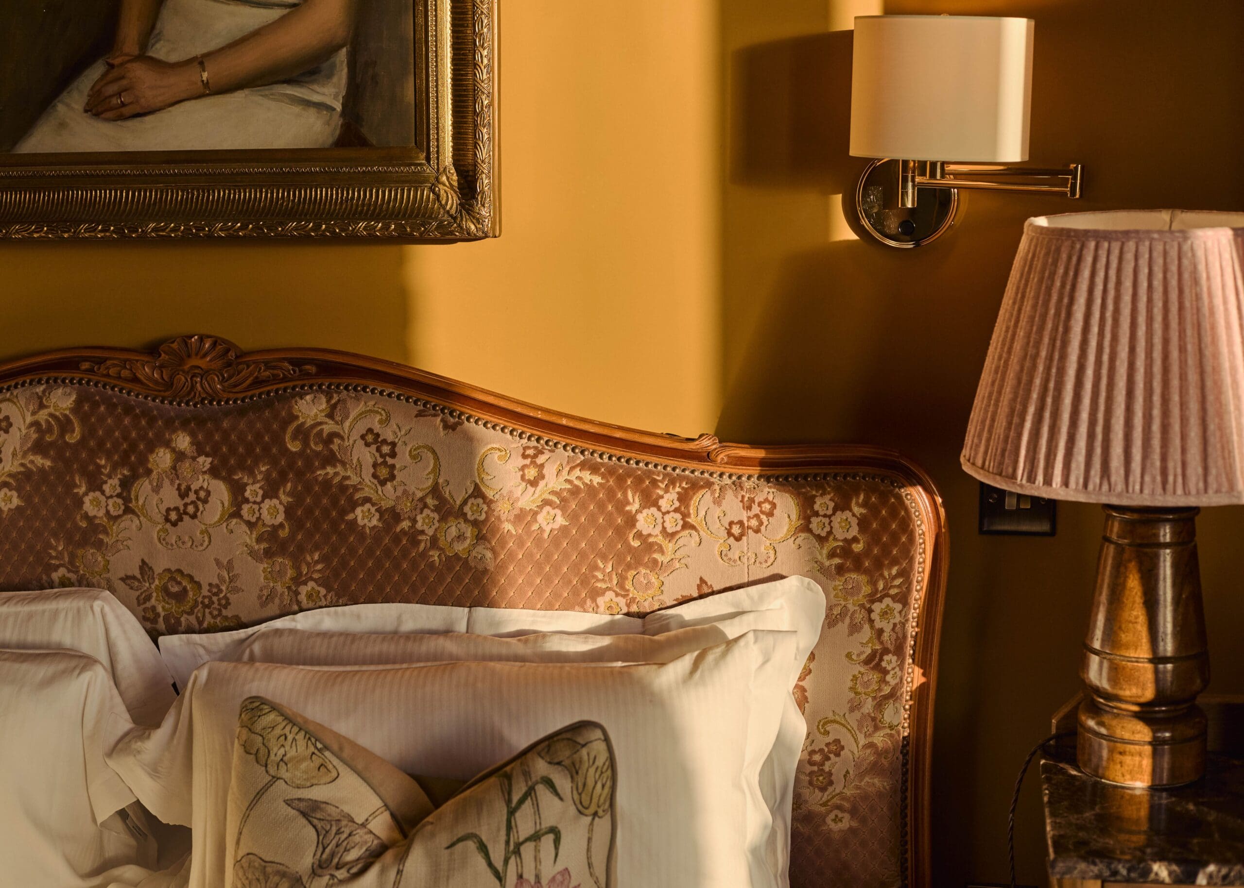 A cozy vintage bedroom with a patterned headboard, white pillows, a floral cushion, a pink pleated lampshade, a wall sconce, and part of a framed portrait in a gold frame, all bathed in warm sunlight.