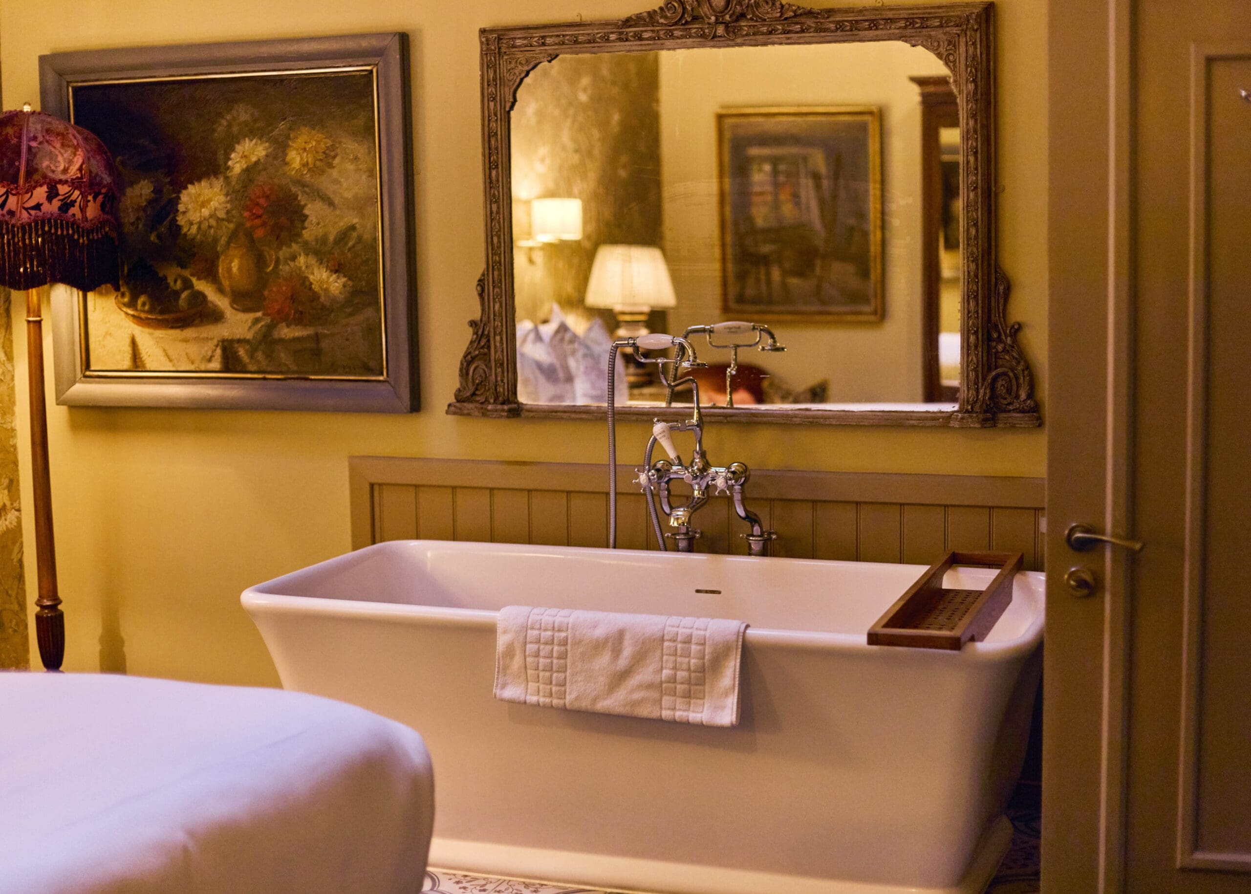A vintage-style bathroom with a white freestanding bathtub, a towel draped over the side, an ornate mirror above, and framed artwork on yellow walls. A lamp and a bed are reflected in the mirror.