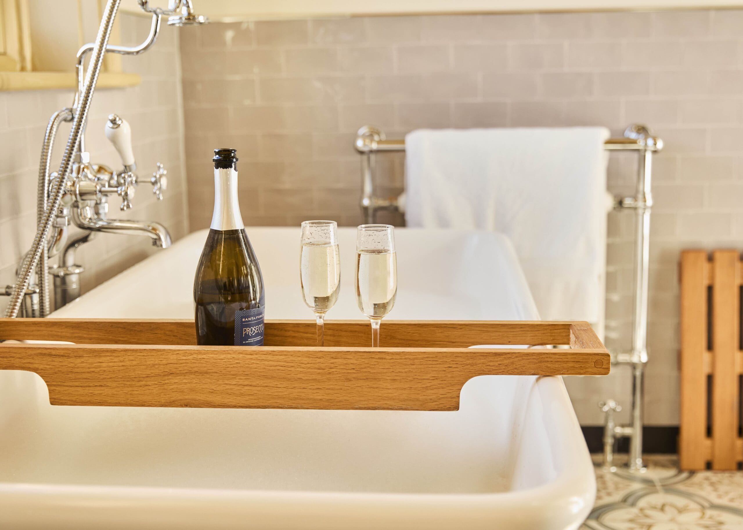 A bottle of sparkling wine and two filled glasses sit on a wooden bath tray across a white bathtub, with a towel on a metal rack and patterned floor tiles in the background.