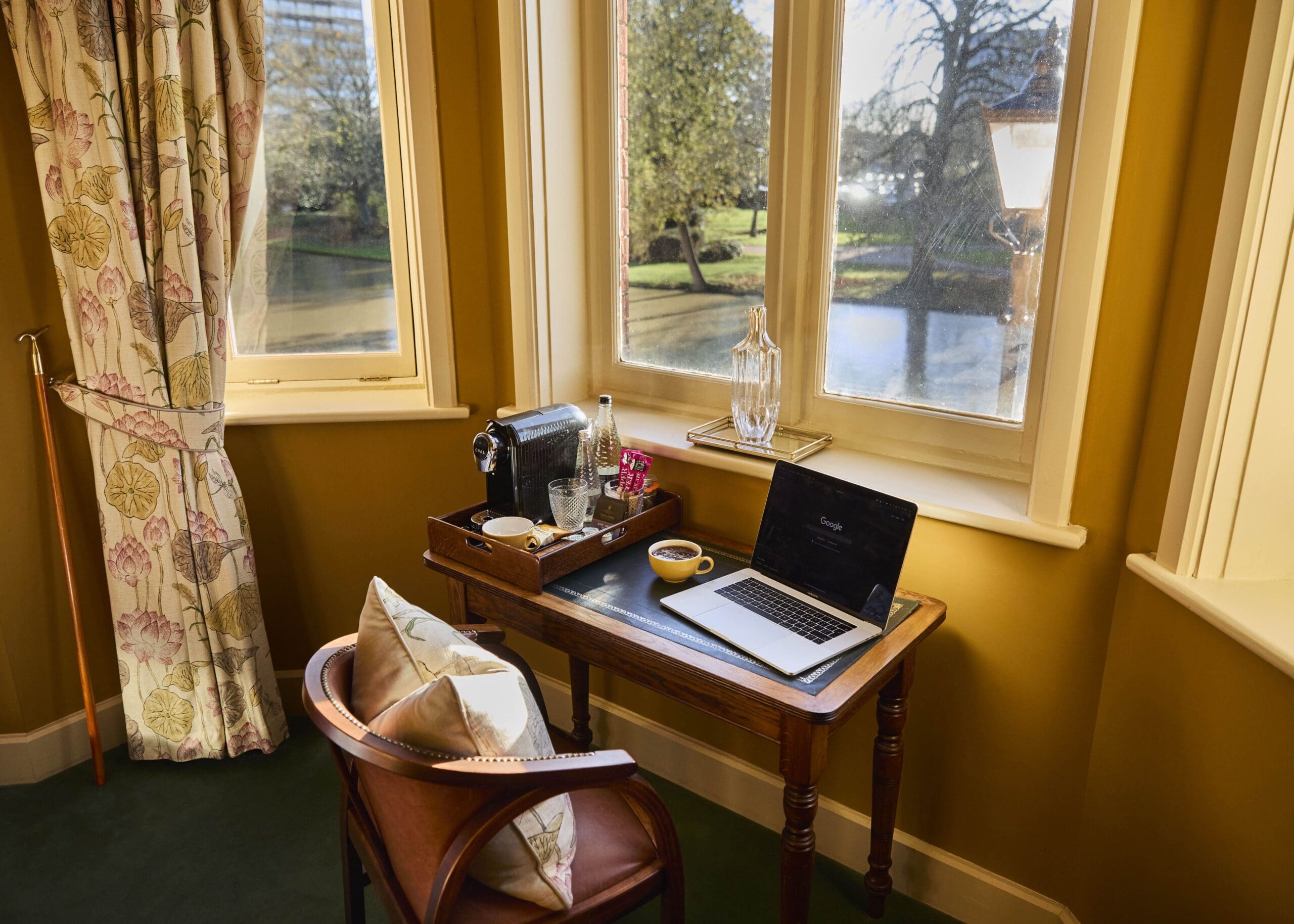 A cozy desk by a window with a laptop, cups, bottled drinks, and glassware on a tray. Sunlight streams through the windows, revealing a scenic view of trees and a river outside. A cushioned chair sits at the desk.