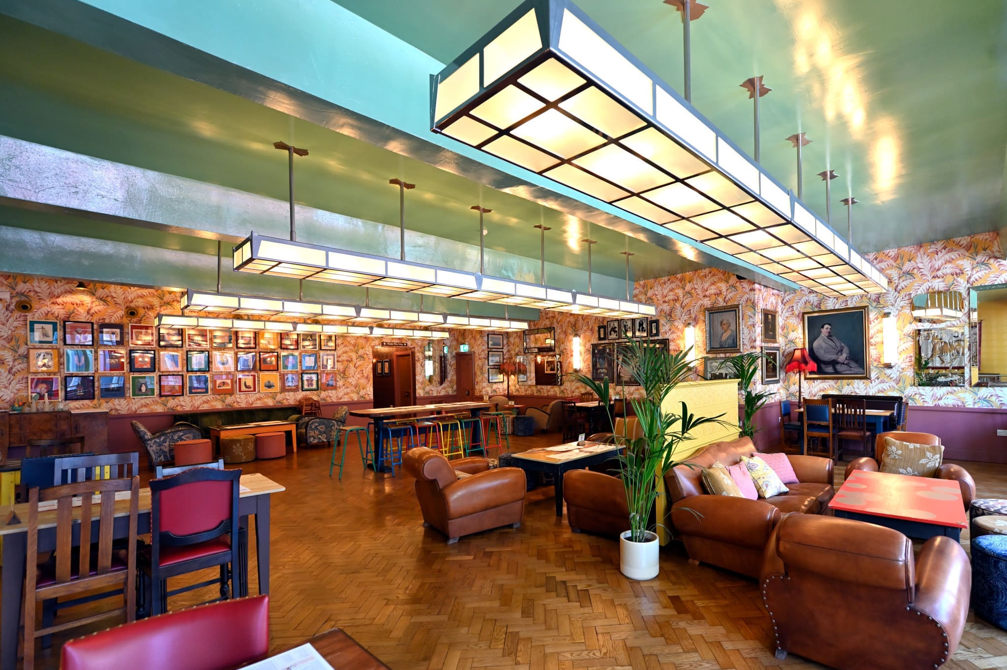 A cozy, eclectic lounge with leather sofas, wooden tables, potted plants, wall art, and portraits. Large rectangular ceiling lights illuminate the space, giving Lupo a welcoming and relaxed atmosphere.