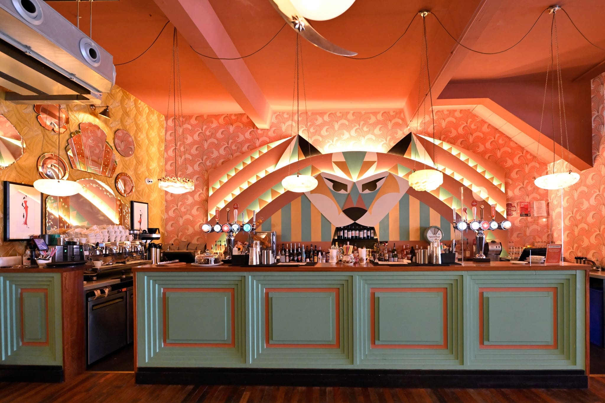 A stylish bar, Lupo, with pastel green paneling, art deco lighting, mirrored wall art, and a bold geometric mural of a stylized face behind the bar, set in a room with warm peach and orange tones.