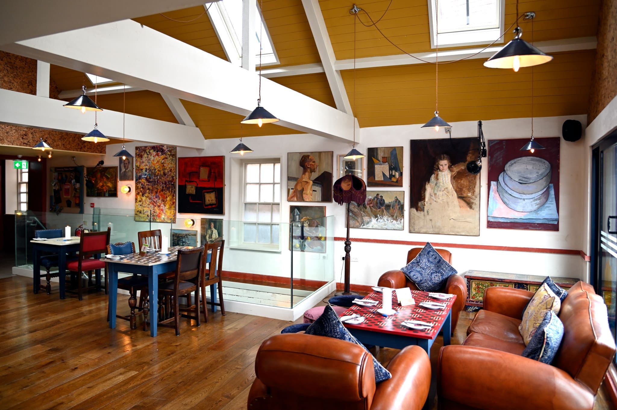 A cozy, art-filled cafe with brown leather sofas, patterned cushions, wooden tables, and hanging pendant lights. Vibrant paintings cover the white and Marisco-yellow walls as sunlight streams in through the windows.