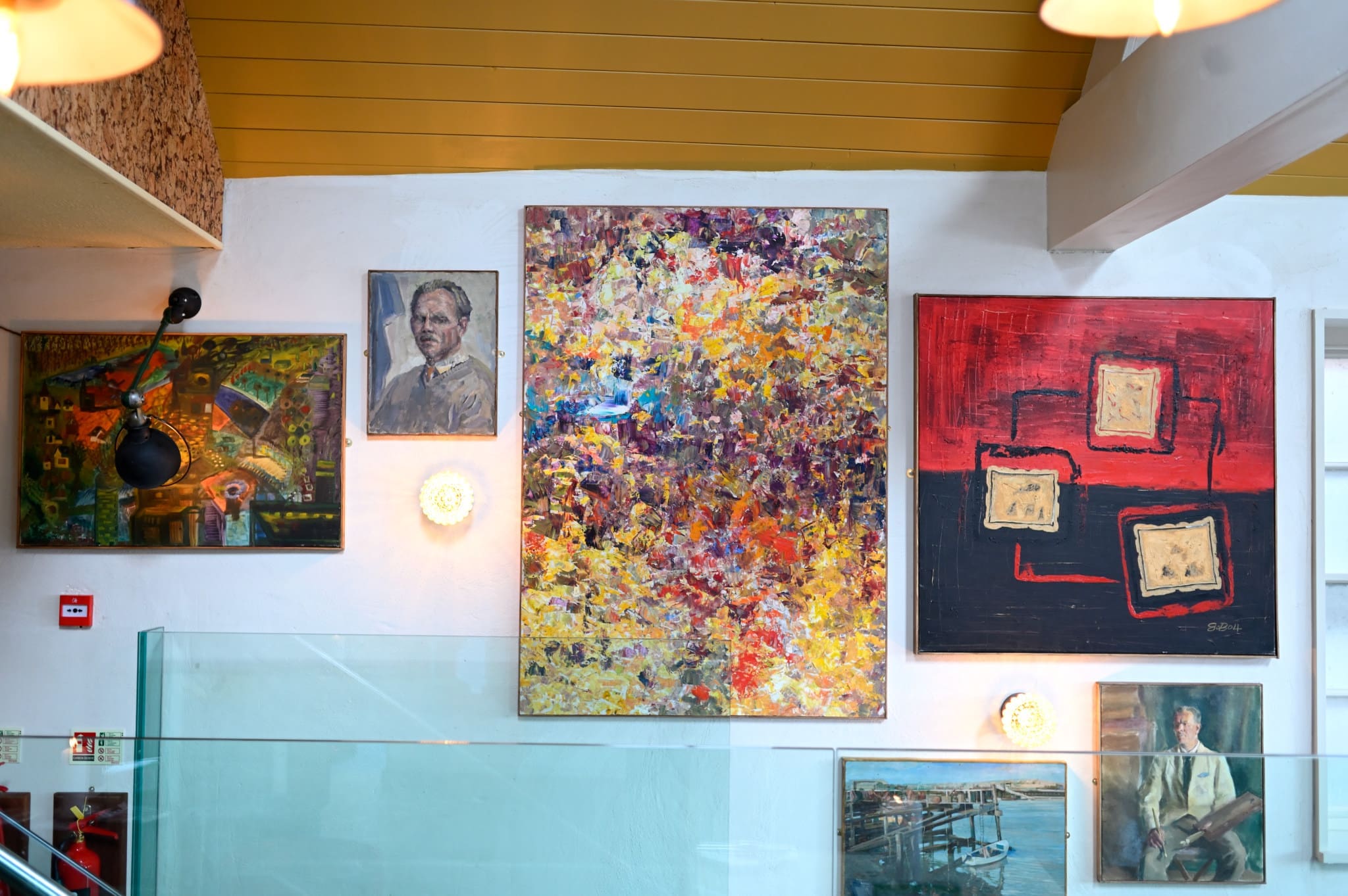 A wall displays various paintings, including Marisco abstract art with vivid colors, portraits, and a red geometric piece, all brightly lit in a modern interior with glass railings and wood accents.