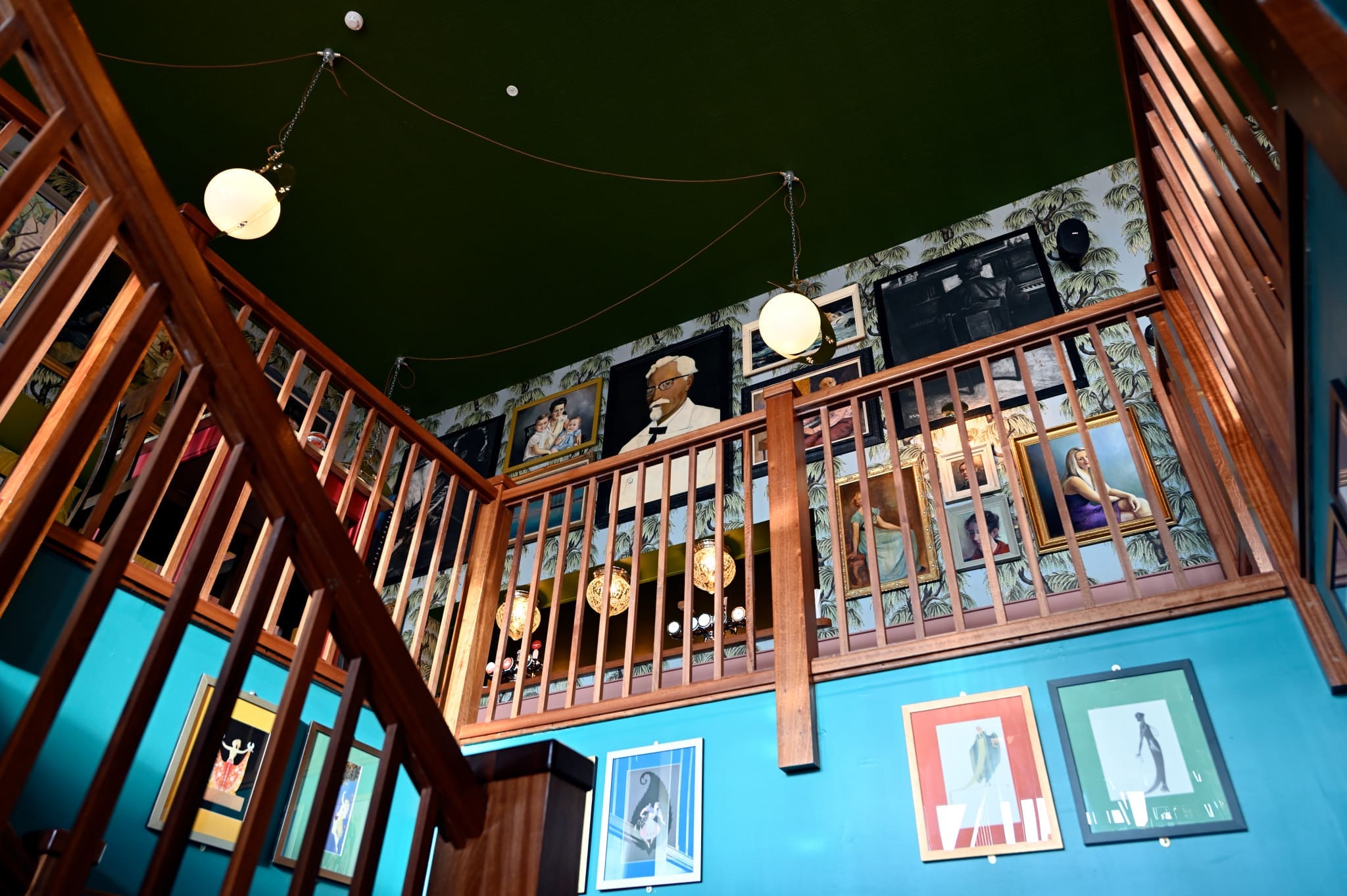 A staircase with Martello wooden railings leads up to a gallery wall decorated with framed portraits and art, tropical wallpaper, and two hanging globe lights under a dark green ceiling.