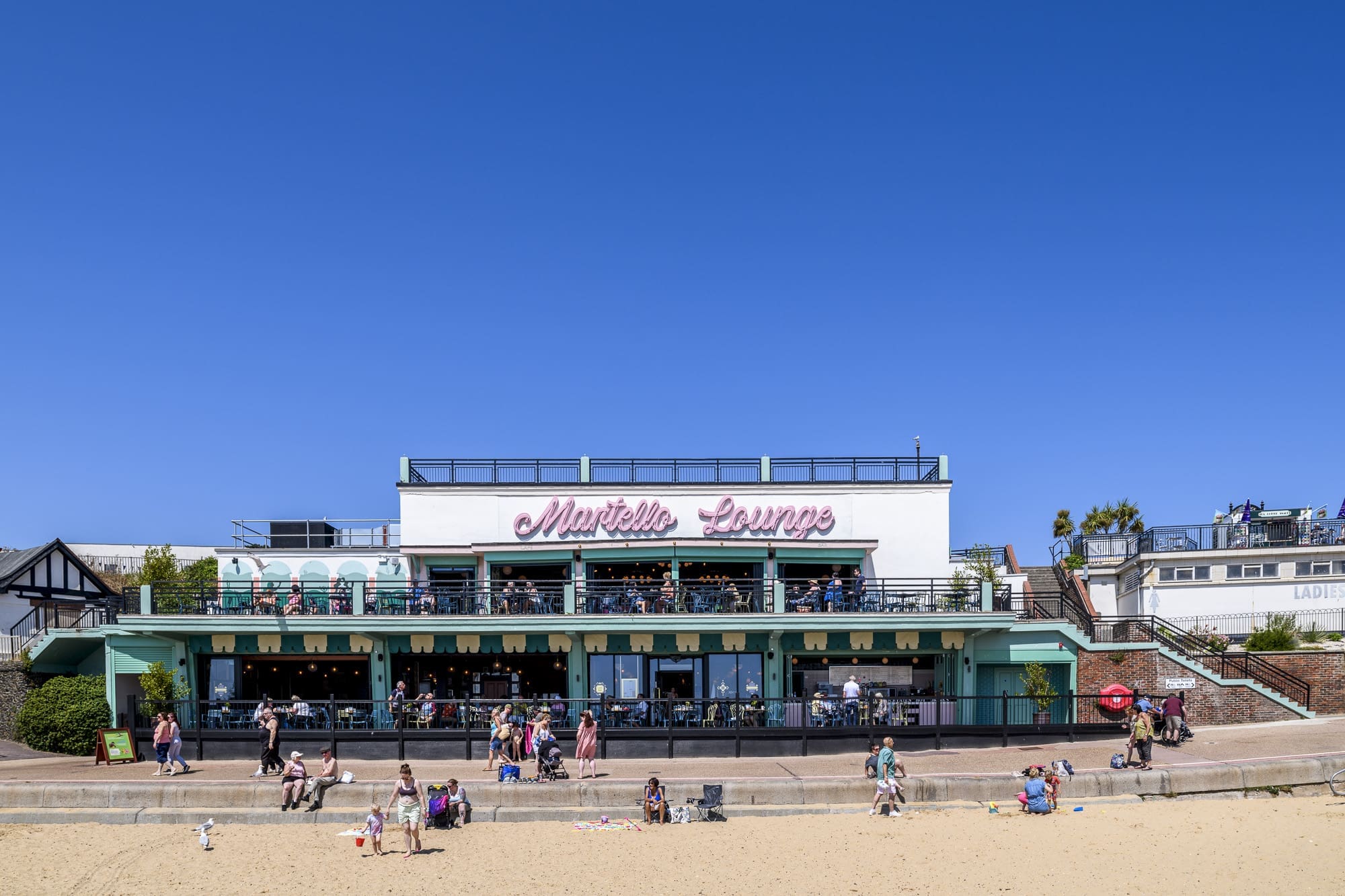 People sit and walk outside Martello Lounge, a beachside restaurant with a pastel green facade, large windows, and an outdoor terrace. The clear blue sky stretches above as visitors relax on the sandy beach in front of Martello.