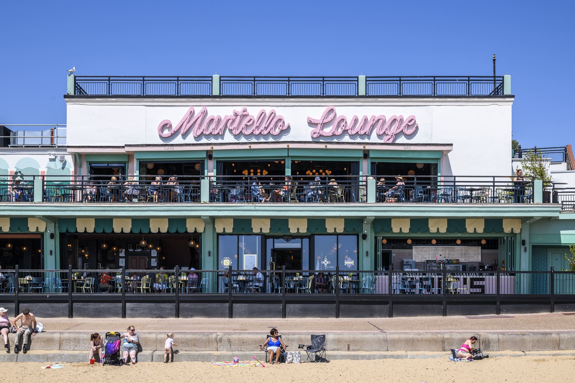 People sit on the sand and promenade in front of the Martello Lounge, a cozy beachside café with pink signage, large windows, and outdoor seating. The clear blue sky adds to the charm of this Martello beachfront spot.