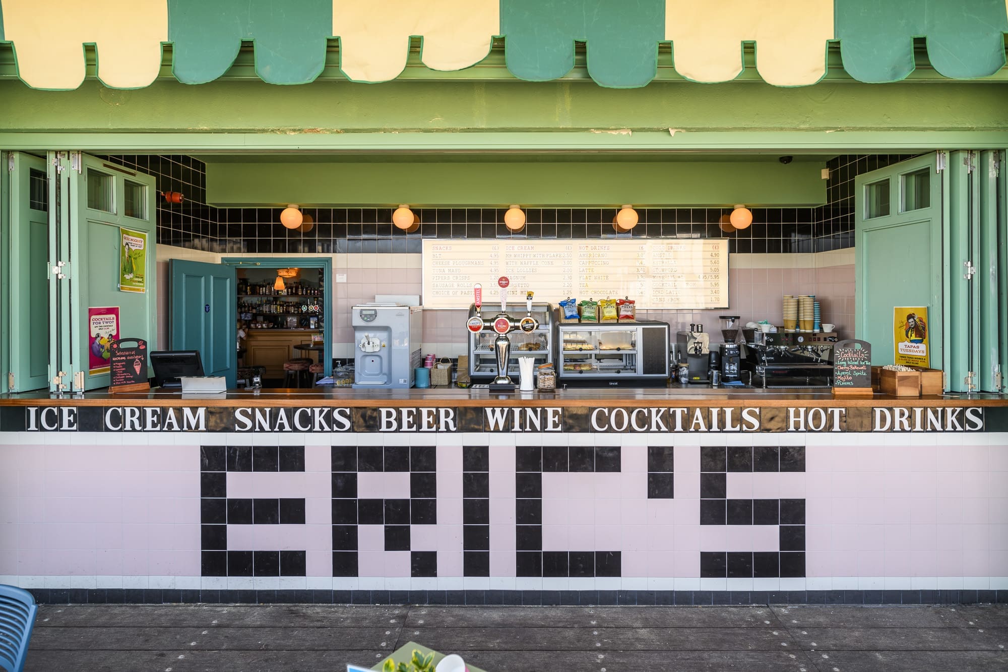 A retro-style snack bar with a green and yellow awning near the Martello, open windows, and counters displaying menus for ice cream, snacks, beer, wine, cocktails, and hot drinks. "ERIC'S" is written in large letters on the front.