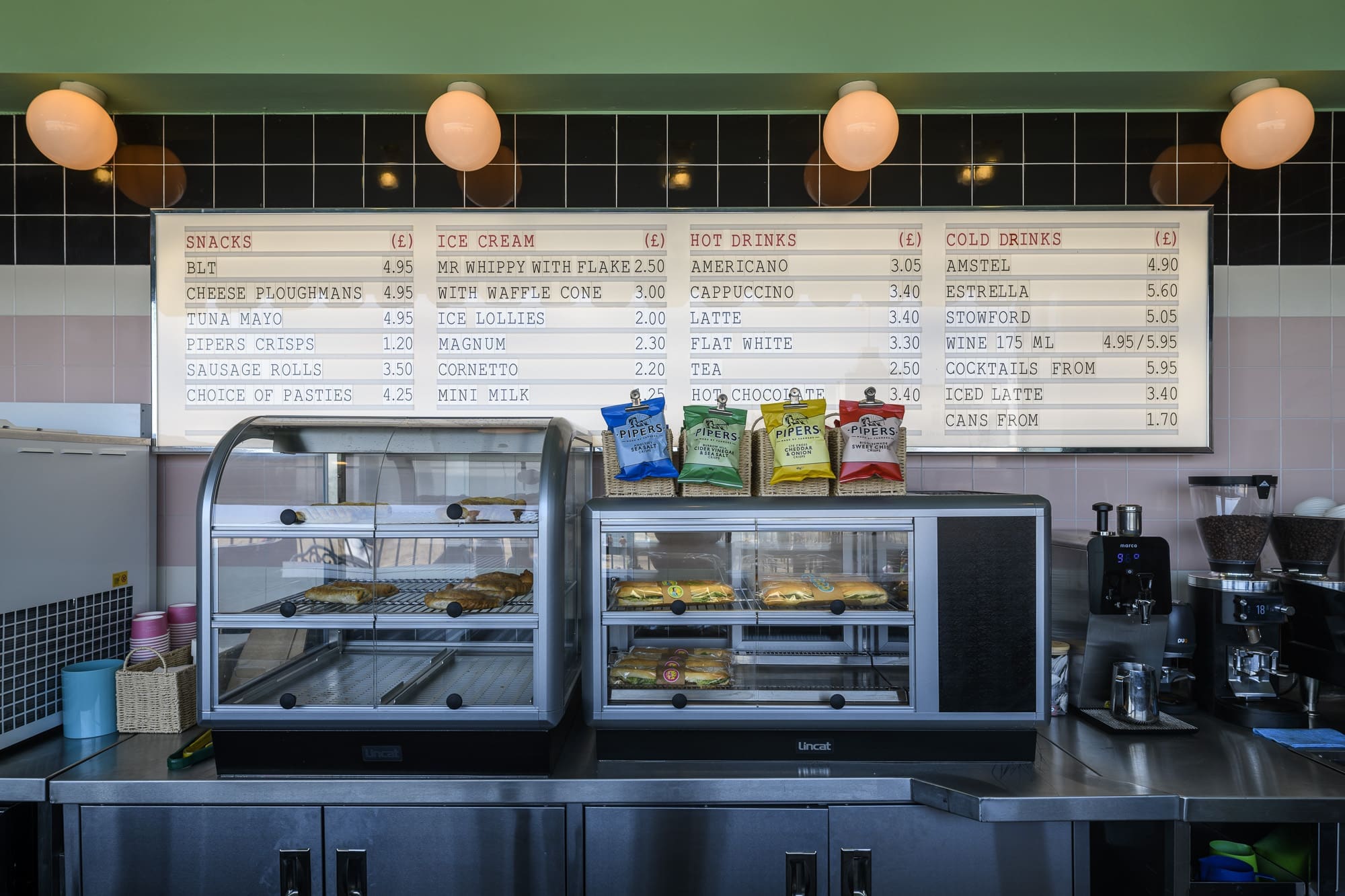 A café counter with two display cases holding pastries and sandwiches, bags of crisps on top, a Martello coffee machine at the side, and a large menu board listing snacks, ice cream, hot drinks, and cold drinks above.