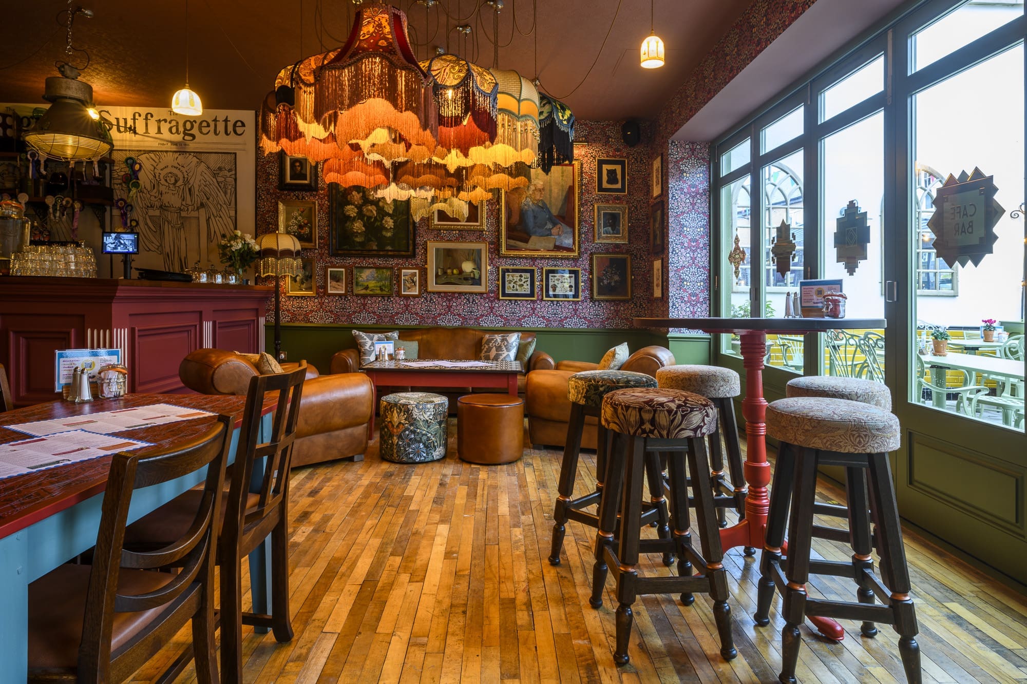 Cozy vintage café interior with patterned wallpaper, eclectic framed art, chandeliers, brown leather sofas, high stools, Martino wooden tables, and large windows overlooking an outdoor patio. Warm ambient lighting.