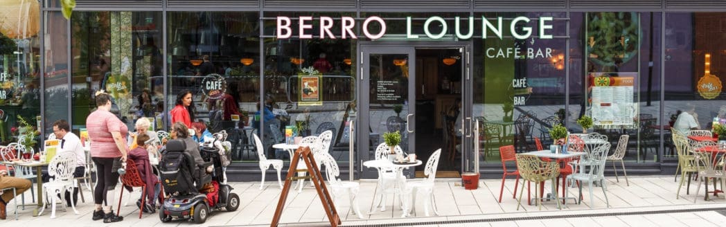 People sit and stand outside Berro Lounge café bar, which has outdoor tables and colorful chairs. The vibrant Berro neon sign glows above large glass windows, and a few people are using mobility aids.