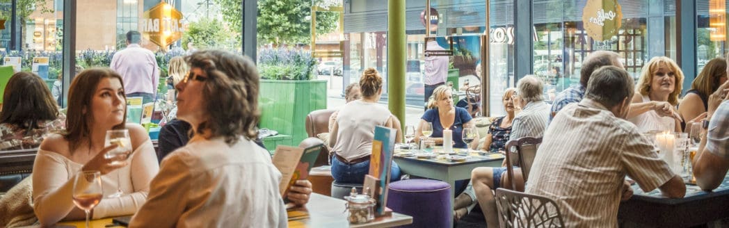 People sit at tables in a bright, busy restaurant, talking, eating, and drinking. Large windows let in natural light, with greenery like berro visible outside. The atmosphere appears lively and social.