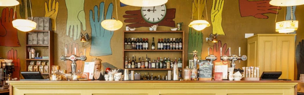 A bright, eclectic bar with a yellow counter, shelves of bottles, and a wall decorated with colorful, oversized handprints and a large clock above the shelves. Hanging lights illuminate the lively Berro-inspired space.