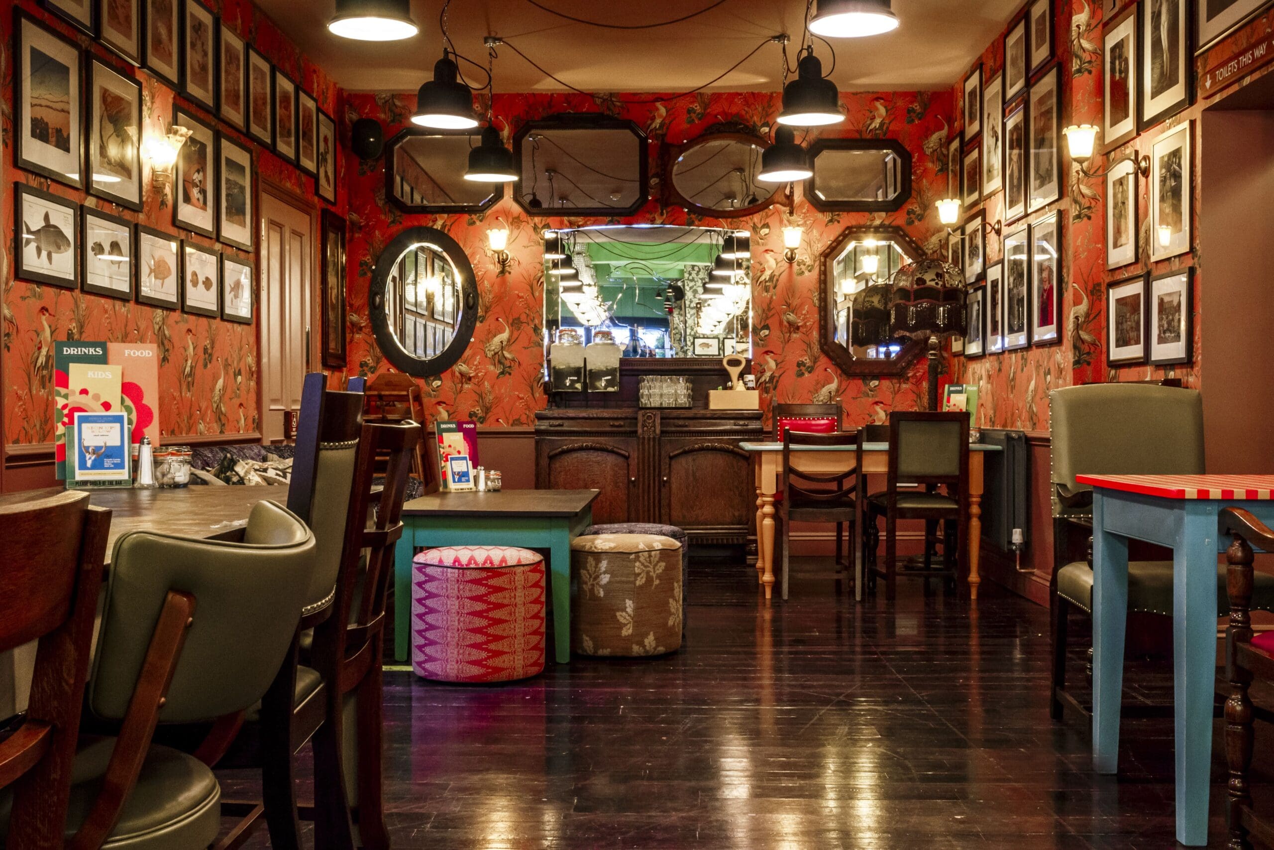 Cozy Granello café interior with eclectic furniture, orange floral wallpaper, framed art on the walls, mirrors, hanging lights, and a polished dark wood floor. The space feels warm and inviting with a vintage, quirky atmosphere.
