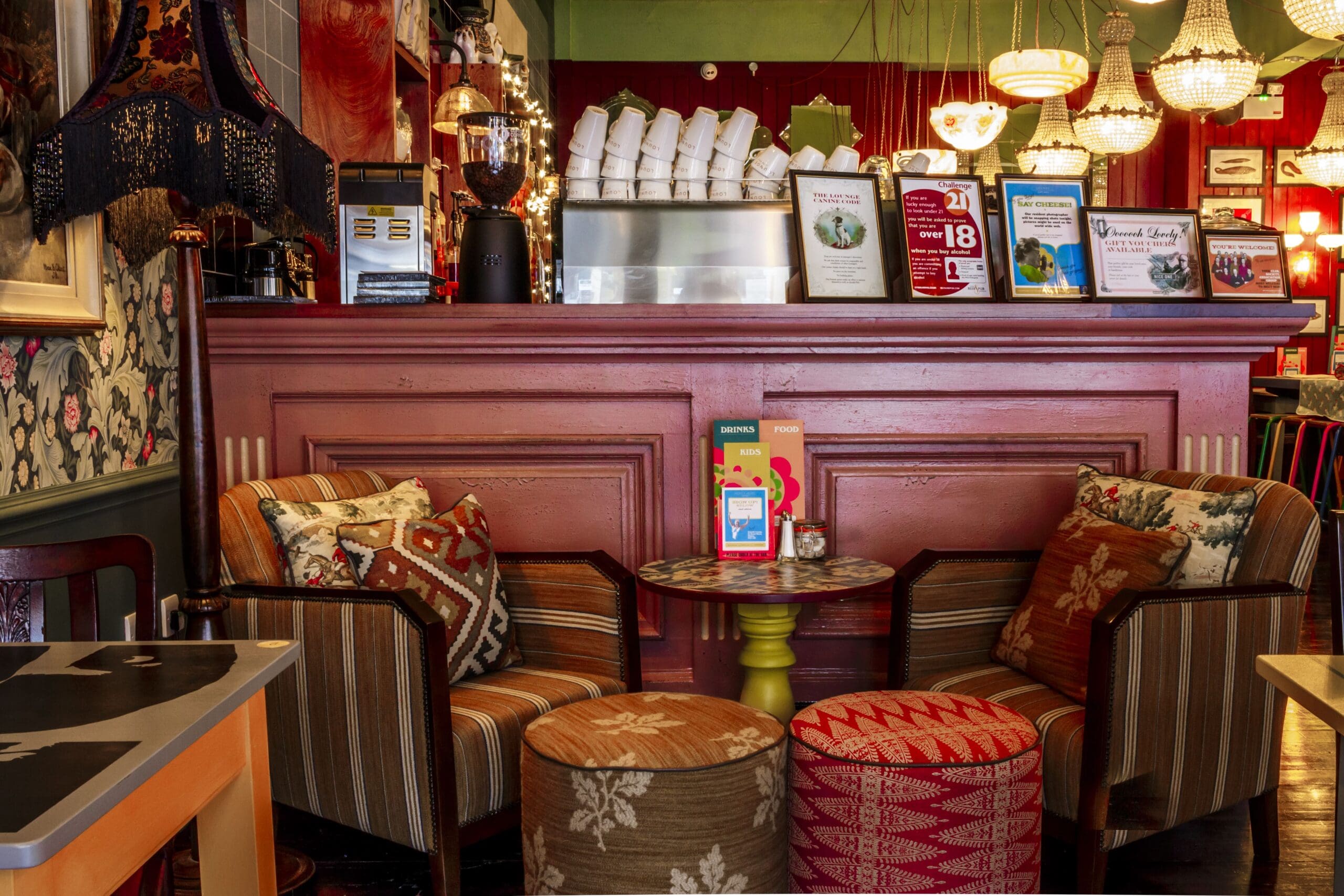 Cozy Granello café scene with two patterned armchairs, round table, and colorful cushions and stools in front of a pink counter. Lamps and chandeliers hang above, with eclectic decor creating a warm, inviting atmosphere.