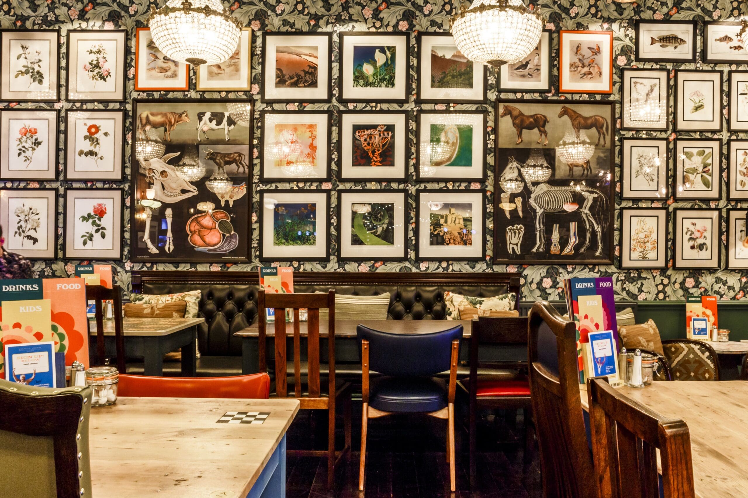 A stylish Granello restaurant interior with wooden tables and chairs, patterned wallpaper, and a wall covered in framed art and animal illustrations. Large chandeliers hang from the ceiling, creating a warm, inviting atmosphere.