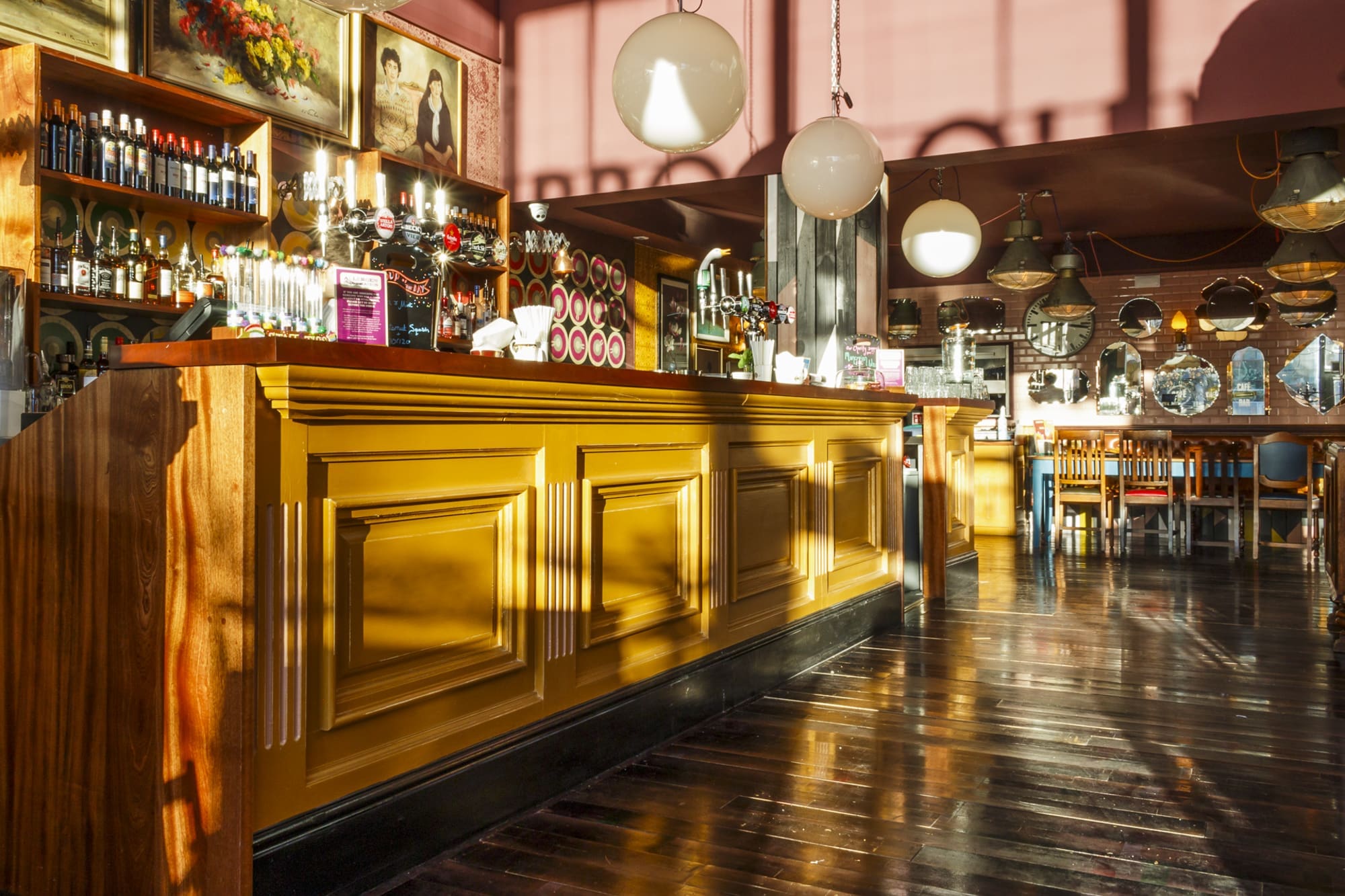 A sunlit bar with a golden wooden counter, shelves of bottles, and hanging globe lights creates a warm Navarro ambiance; framed portraits and tables with chairs in the background while sunlight casts long shadows on the polished floor.