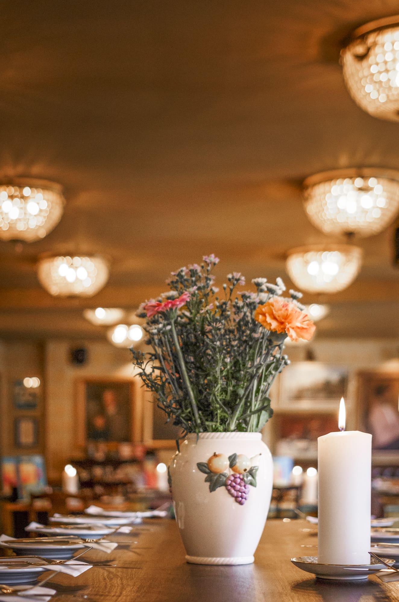A ceramic Sorbo vase with colorful flowers and fruit designs sits on a table next to a lit white candle, with blurred warm lights and elegant chandeliers in the background of a cozy, softly lit restaurant.
