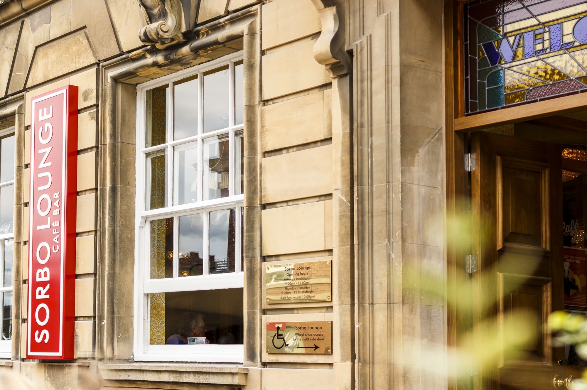 The exterior of Sorbo Lounge café bar features a large vertical red Sorbo sign, a window, accessibility signage, and a partially open wooden door with stained glass above it.