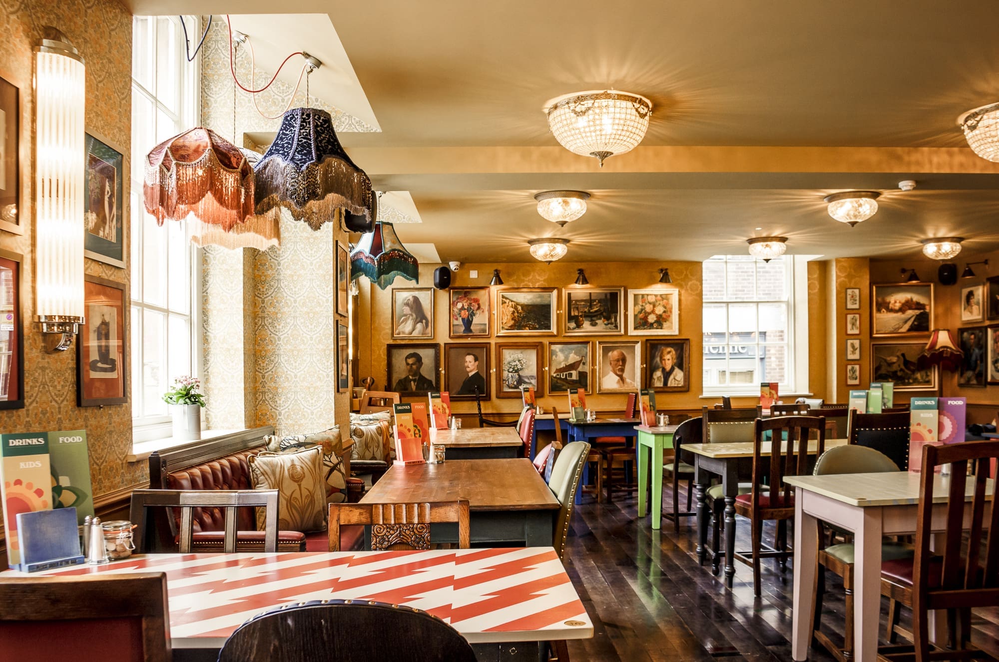 A brightly lit, eclectic café with vintage decor and Sorbo charm features mismatched chairs and tables, colorful lampshades, and walls covered in assorted framed portraits and art. Large windows flood the space with natural light.