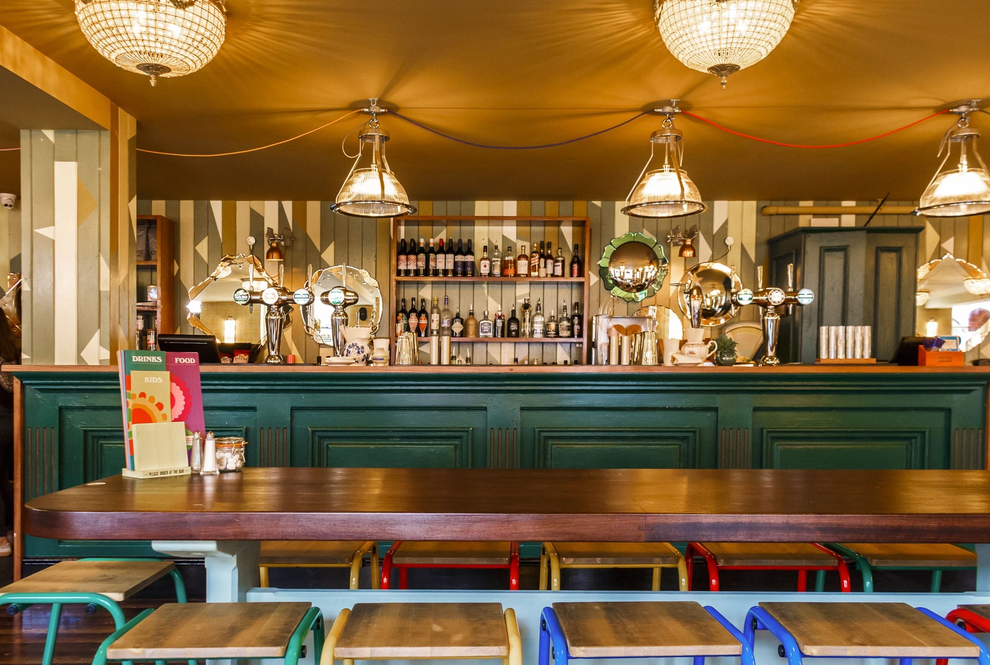 A cozy, stylish bar interior featuring a long wooden table, colorful stools, and a green Sorbo bar counter, with shelves of bottles and hanging vintage lights glowing under a warm yellow ceiling.