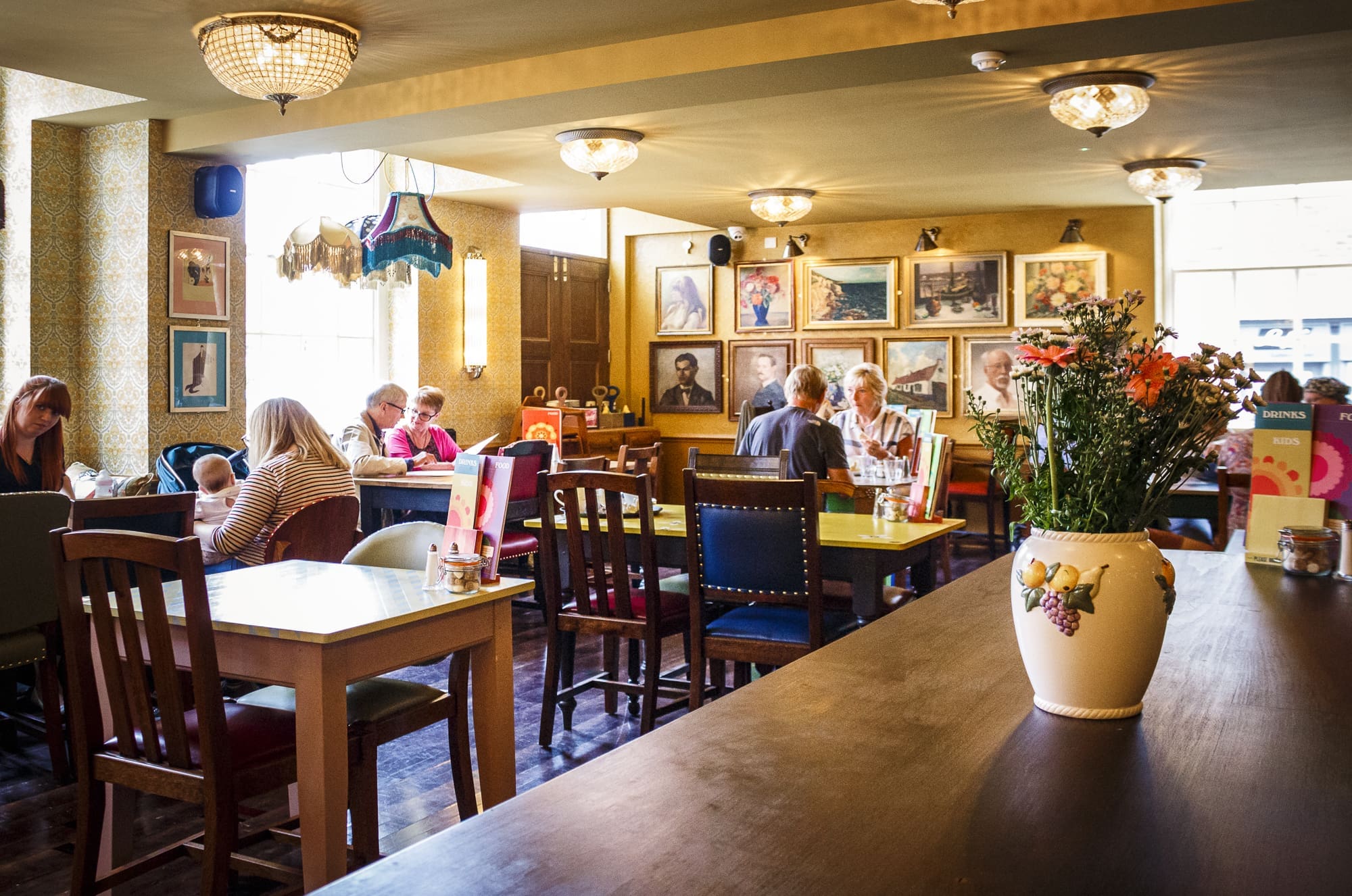 A cozy restaurant interior with several groups dining at wooden tables. Sorbo decor accents the walls with framed pictures, while a vase of flowers sits on a counter in the foreground. Warm lighting creates a welcoming atmosphere.
