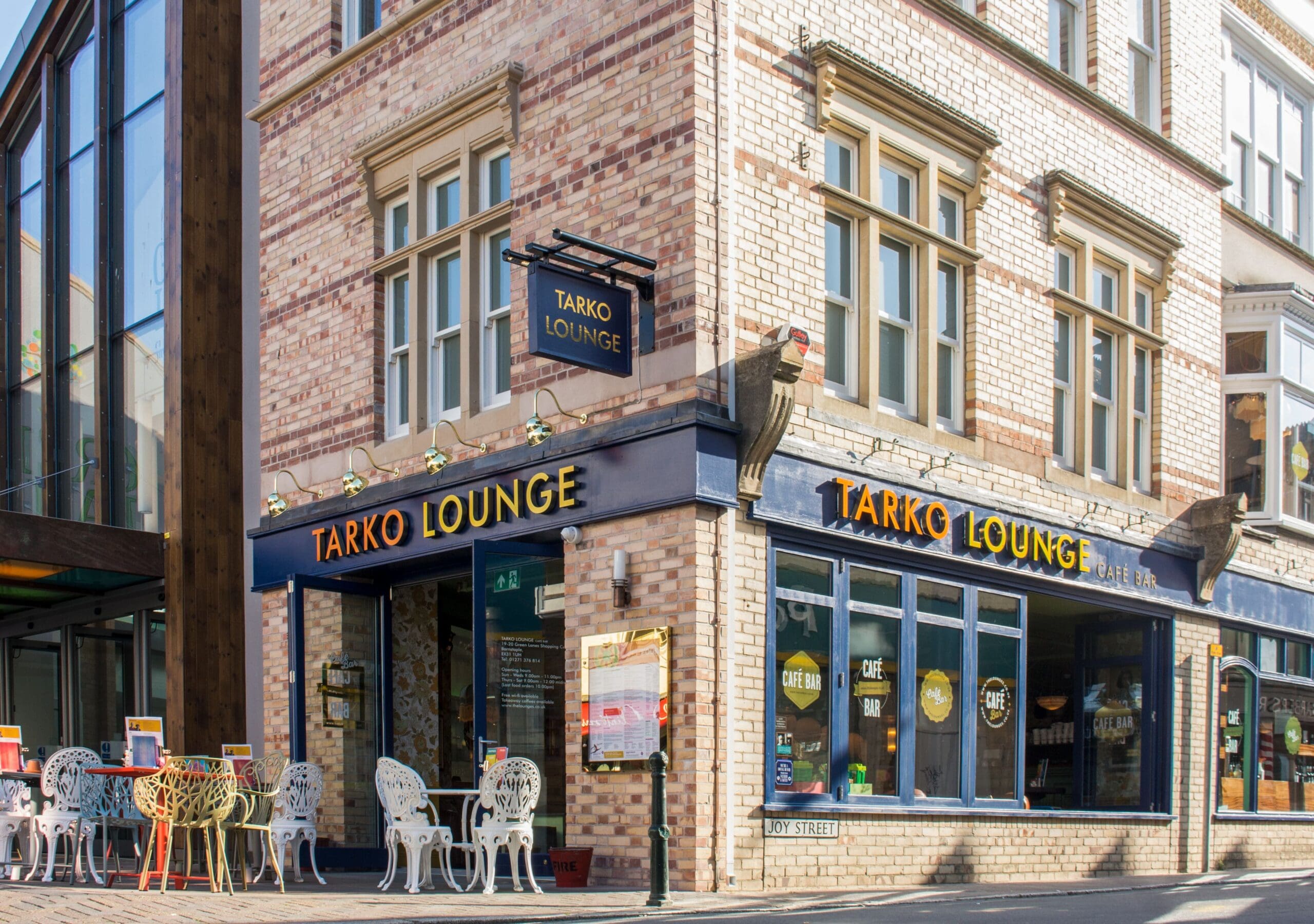 A corner café called "Tarko Lounge" with large windows, outdoor tables and chairs, and a brick exterior basks in the sunlight. The street sign reads "Judy Street." Tarko’s inviting vibe draws passersby to relax and enjoy the day.