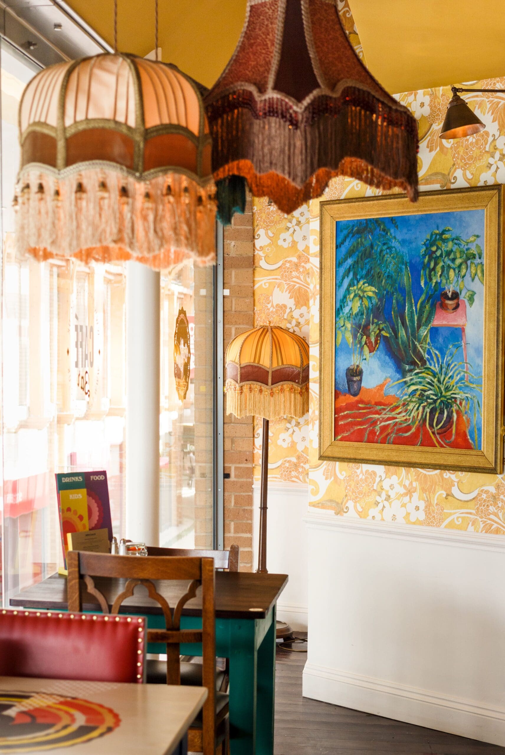 A cozy Tarko cafe interior with vintage hanging lamps, patterned wallpaper, a framed painting of plants, a retro floor lamp, and a small table with chairs by the window.