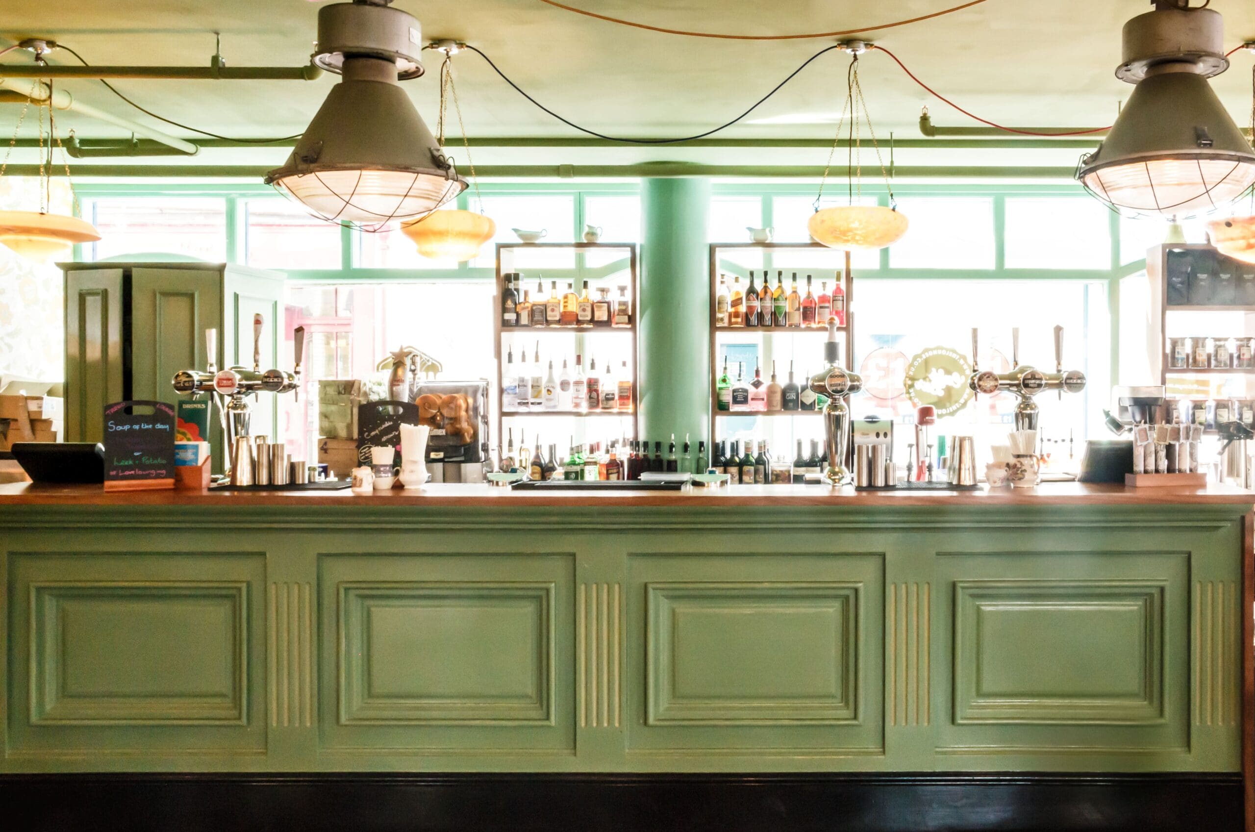 A brightly lit Tarko bar with a pastel green counter, shelves stocked with bottles of alcohol, hanging industrial-style lights, and large windows letting in natural light.