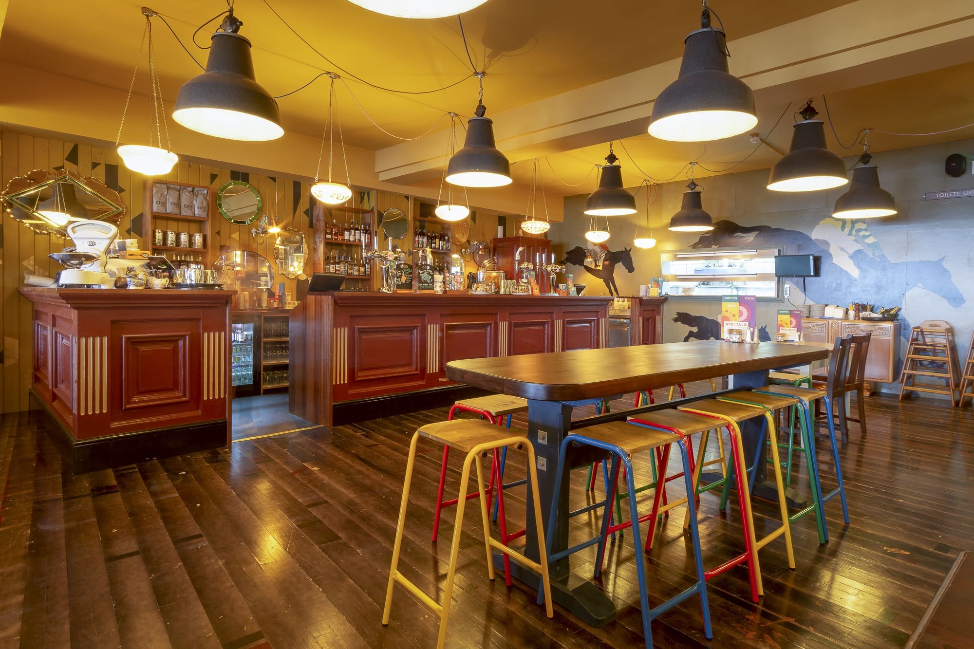 A cozy café interior with warm lighting, wooden floors, a long wooden table with colorful stools, and an inviting Orsino bar counter lined with various bottles and coffee equipment in the background.