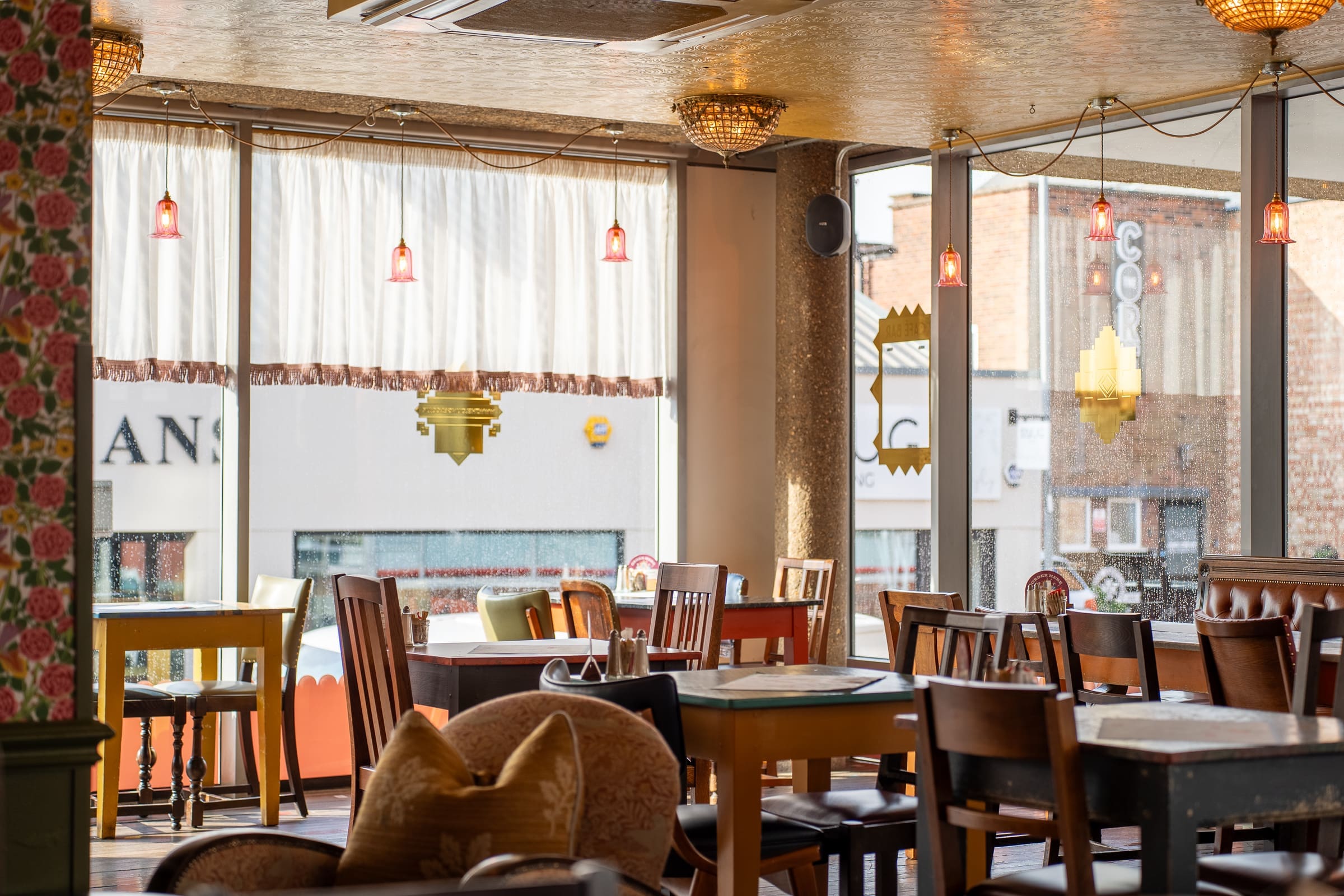 A cozy Mercado restaurant interior with wooden tables and chairs, warm lighting from hanging lamps, and large windows with white curtains letting in natural light.