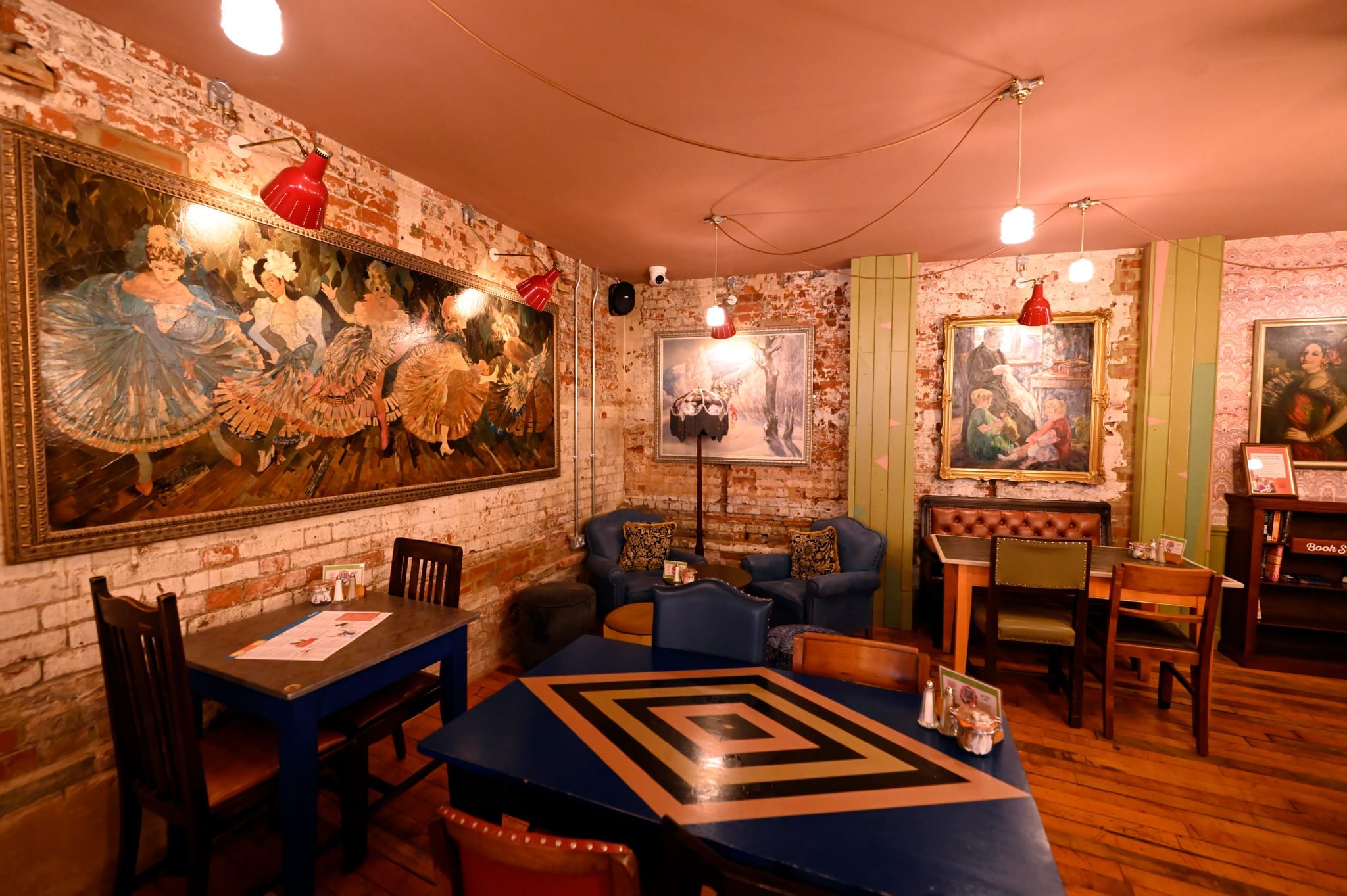 Cozy cafe with exposed brick walls, featuring framed art, eclectic Merletto-inspired furniture including tables, booths, and armchairs, and warm lighting from hanging bulbs and red wall lamps.