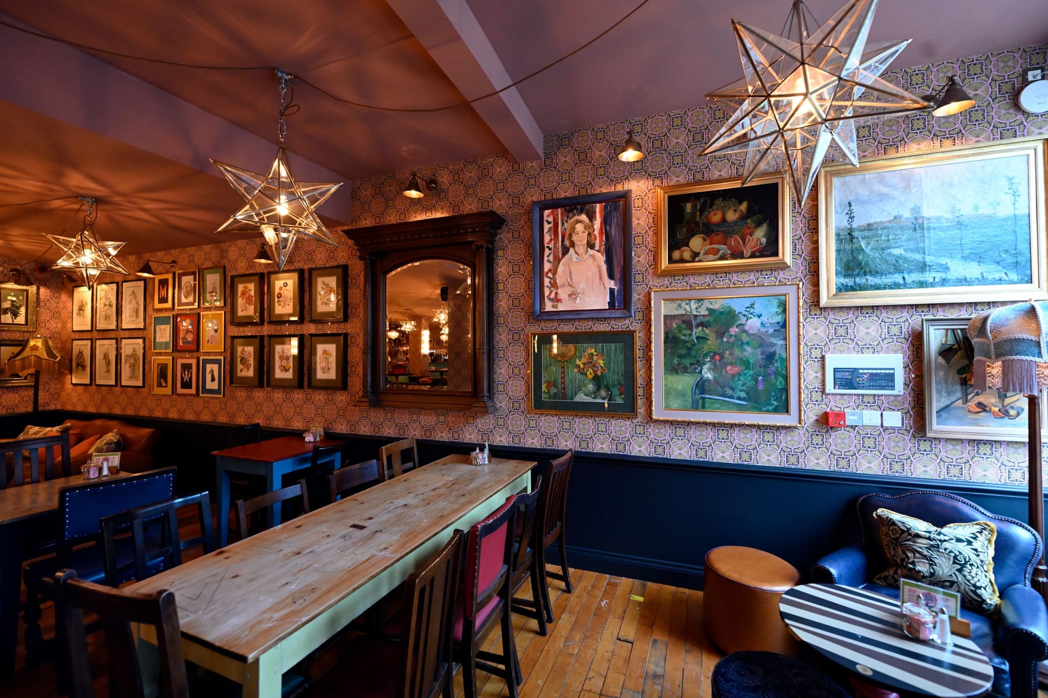 A cozy café interior with wooden tables, eclectic chairs, star-shaped pendant lights, and Merletto-inspired hardwood floors. Walls adorned with framed paintings and photographs enhance the warm and inviting atmosphere.