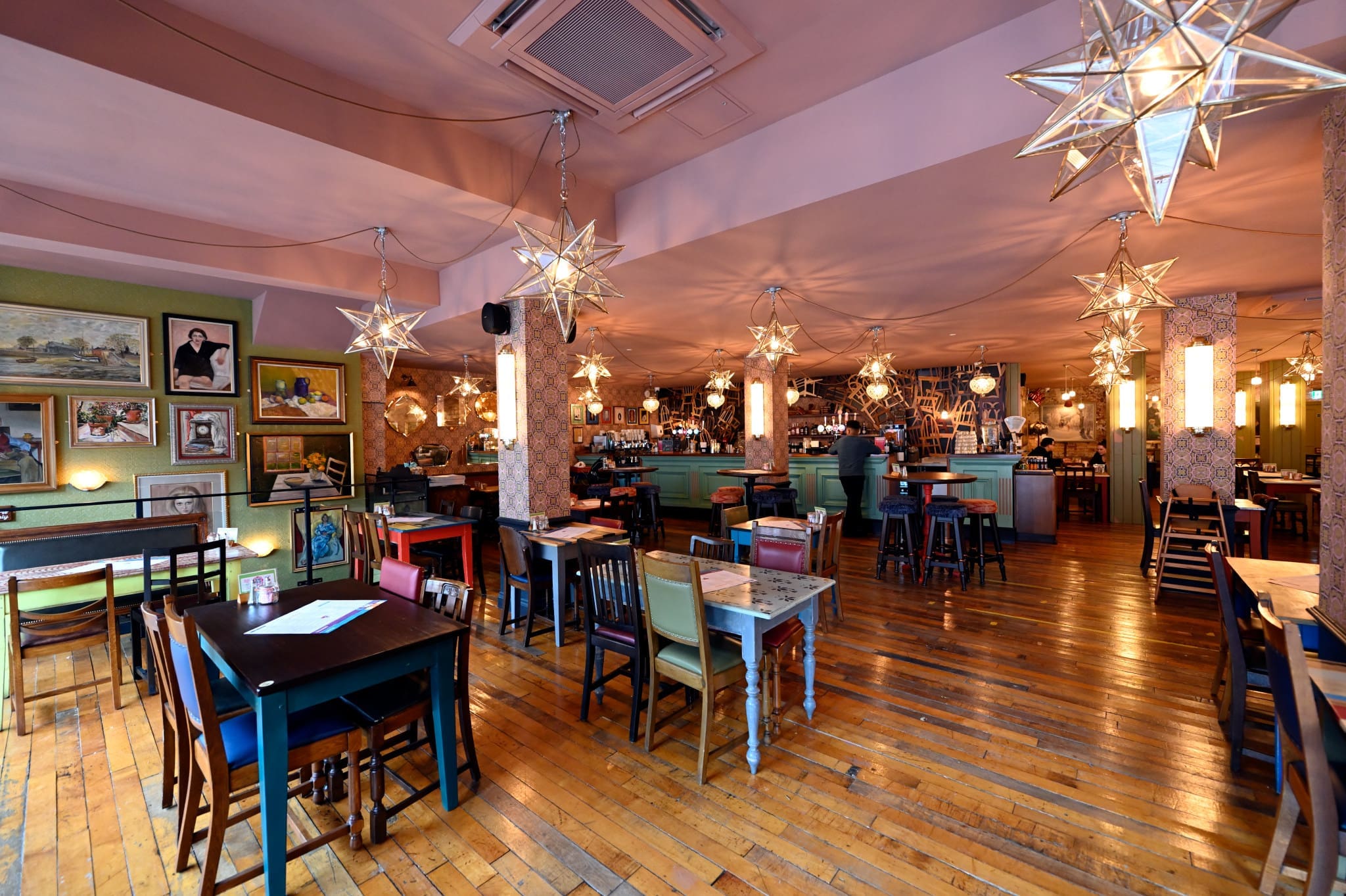 A cozy Merletto restaurant with wooden floors, eclectic tables and chairs, star-shaped hanging lights, and framed artwork on the walls, with a welcoming bar area visible in the background.