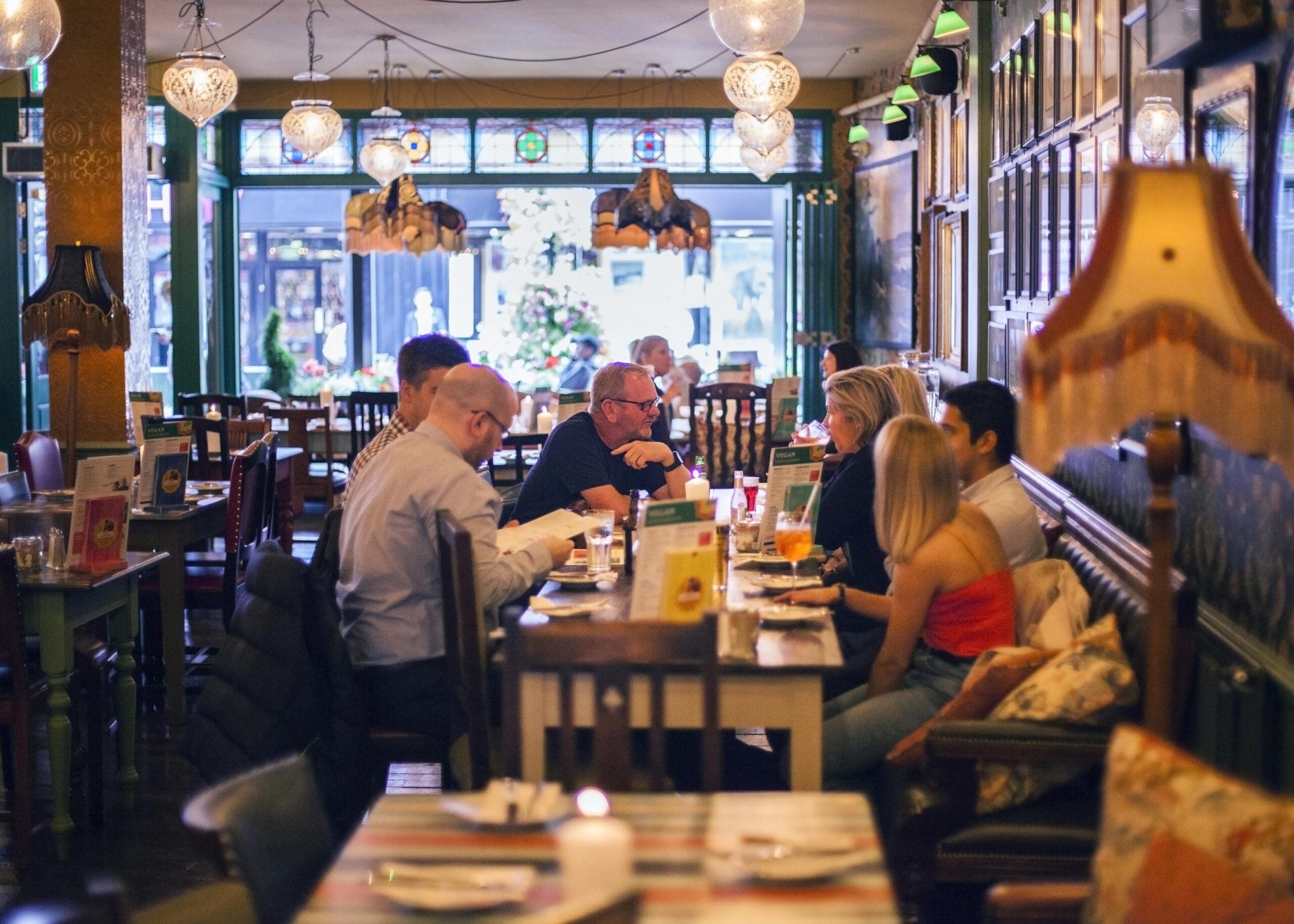 A group of people sit at a long table in Poco, a cozy, warmly-lit restaurant with vintage decor, enjoying food and conversation. Other tables, lamps, and large windows are visible in the background.