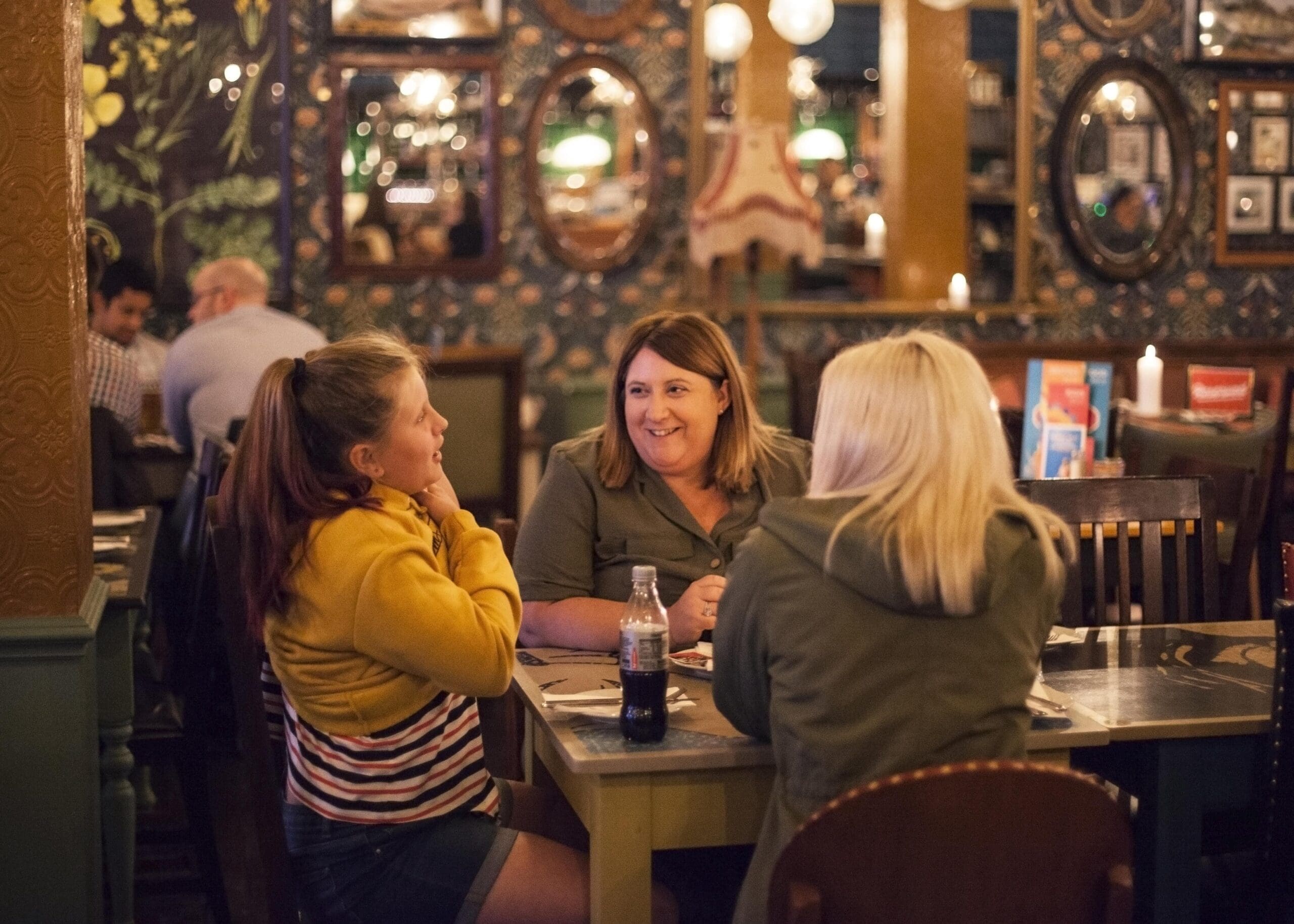 Three women sit and chat at a warmly lit Poco restaurant table, surrounded by ornate wallpaper, framed mirrors, and decorative lights, creating a cozy and inviting atmosphere.