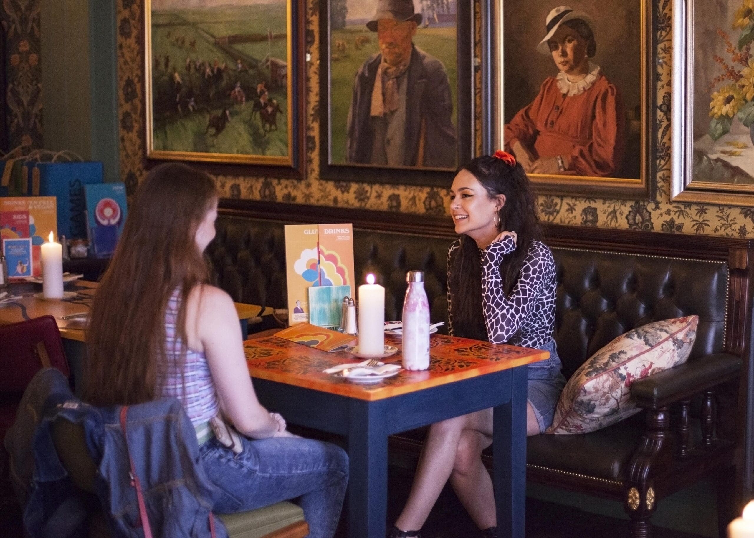 Two women sit across from each other at a candlelit table in Poco, a cozy, art-filled restaurant, smiling and engaged in conversation. Paintings and colorful menus decorate the warmly lit space.