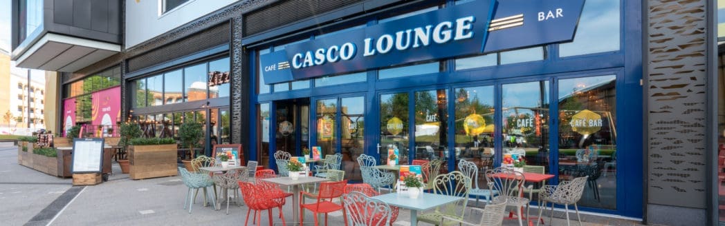Outdoor seating area with colorful chairs and tables in front of Casco Lounge café and bar. Large glass windows reflect nearby buildings and trees, while Casco’s vibrant plants and decorations can be seen inside and outside the café.