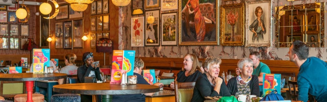 A group of people sit at tables in a cozy, art-filled restaurant in Casco, chatting and smiling. Colorful menus are on the tables, and framed artwork and mirrors decorate the walls. Warm lighting gives the space a lively, inviting feel.