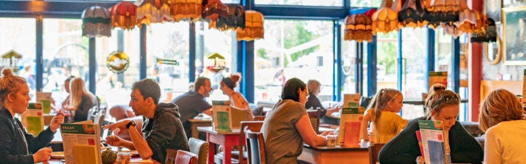 People sit at tables eating and reading menus inside Casco, a brightly lit restaurant with colorful, hanging stained-glass lamps and large windows letting in natural light.