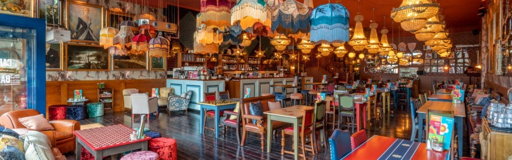 A colorful, eclectic Casco restaurant interior with mismatched chairs and tables, vibrant lampshades hanging from the ceiling, chandeliers, framed art on the walls, and a cozy, inviting atmosphere.