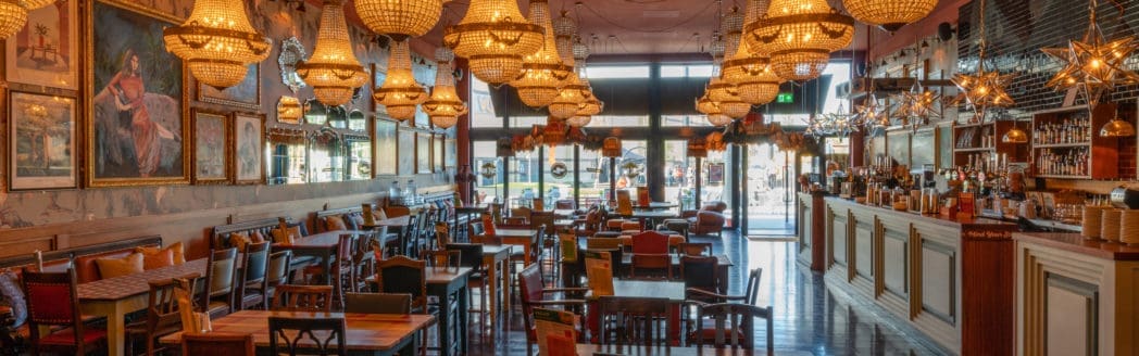 A spacious, elegant restaurant interior in Casco with ornate chandeliers, eclectic paintings on the wall, a variety of tables and chairs, and a well-stocked bar on the right. Natural light streams in through large front windows.
