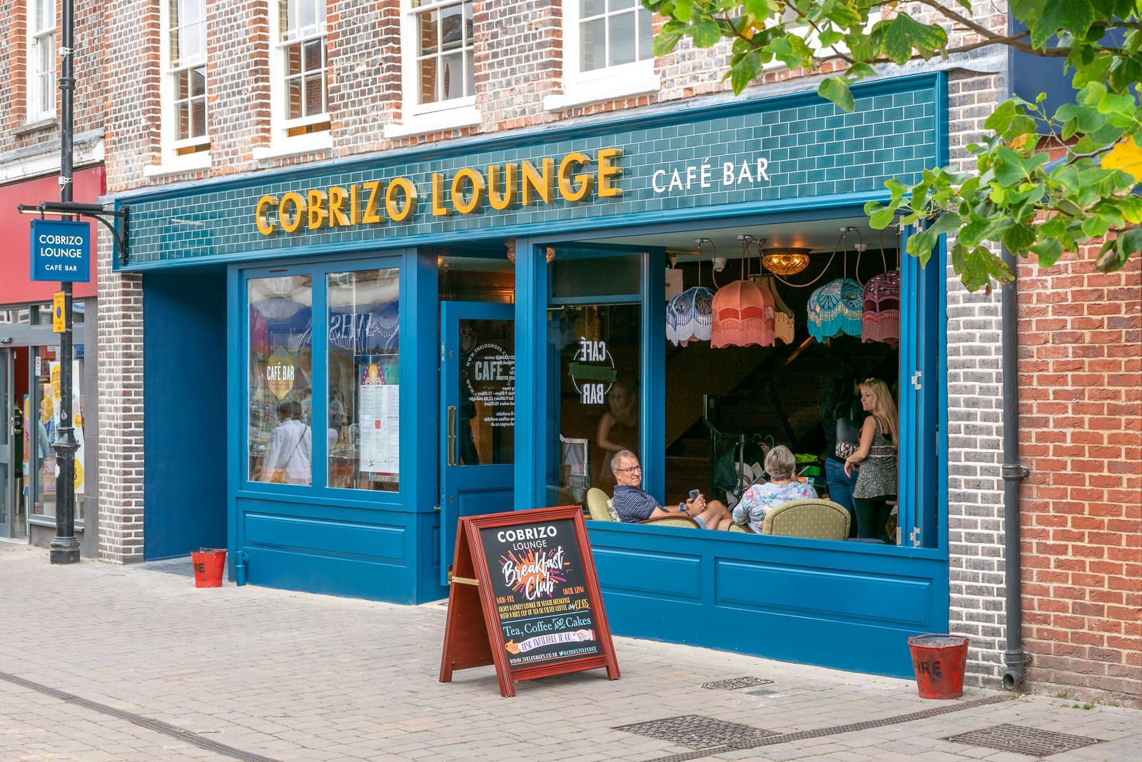 Front view of Cobrizo Lounge café bar with large blue windows, a sidewalk chalkboard sign, and people sitting inside. The Cobrizo exterior features teal tiles and exposed brick for a welcoming vibe.