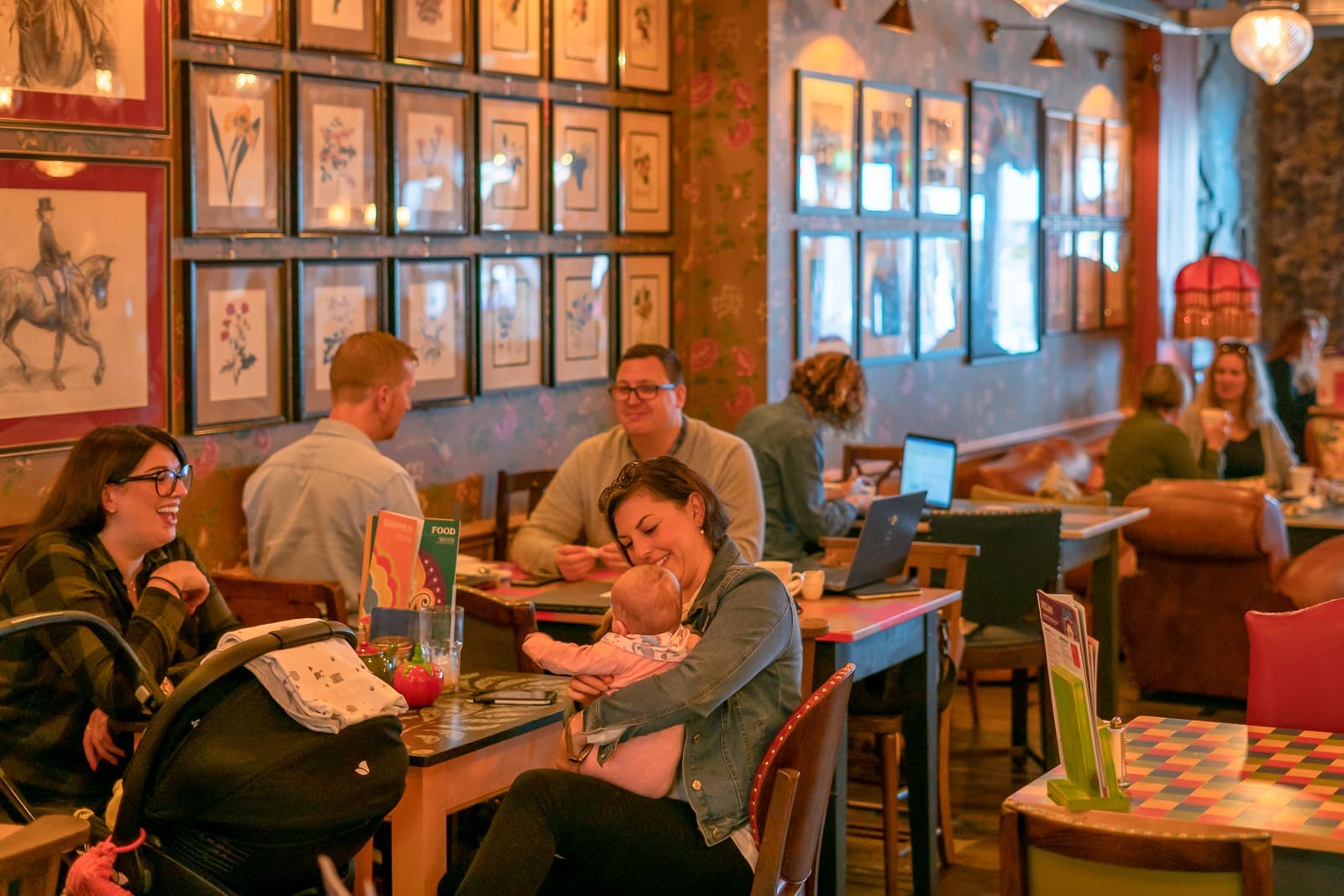 A lively café with people sitting at tables, chatting, working on laptops, and a woman smiling while holding a baby. Framed artwork and cobrizo accents cover the walls, creating a cozy, social atmosphere.