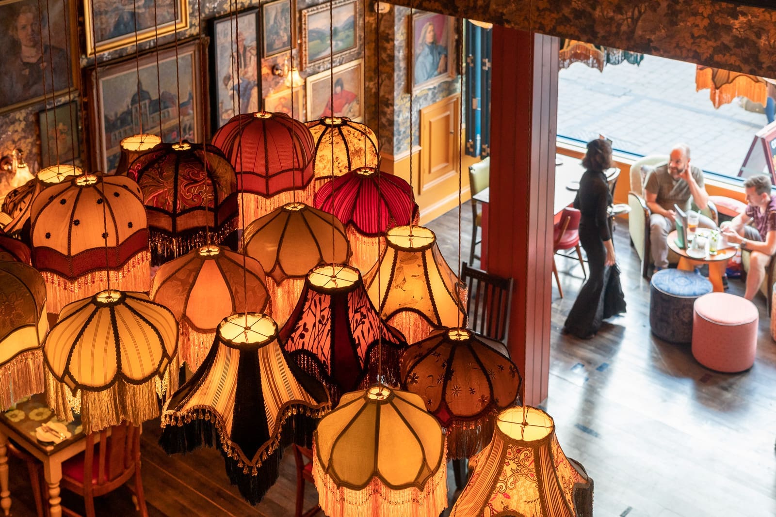 A cluster of cobrizo vintage lampshades hangs from the ceiling in a warmly lit café, with framed pictures on the walls and three people conversing at a table near a large window.