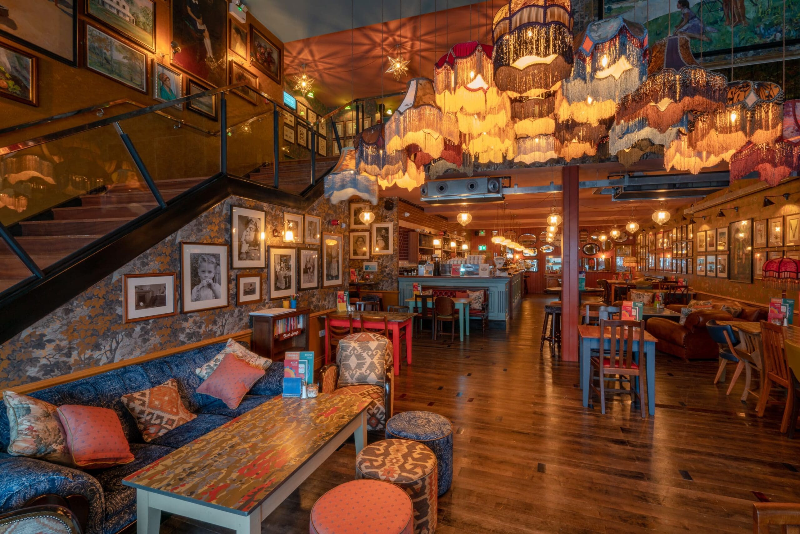 A cozy, eclectic restaurant interior with vintage lampshades hanging from the ceiling, colorful furniture, and framed photos on the walls. Warm cobrizo lighting creates an inviting atmosphere as a staircase leads to an upper level.