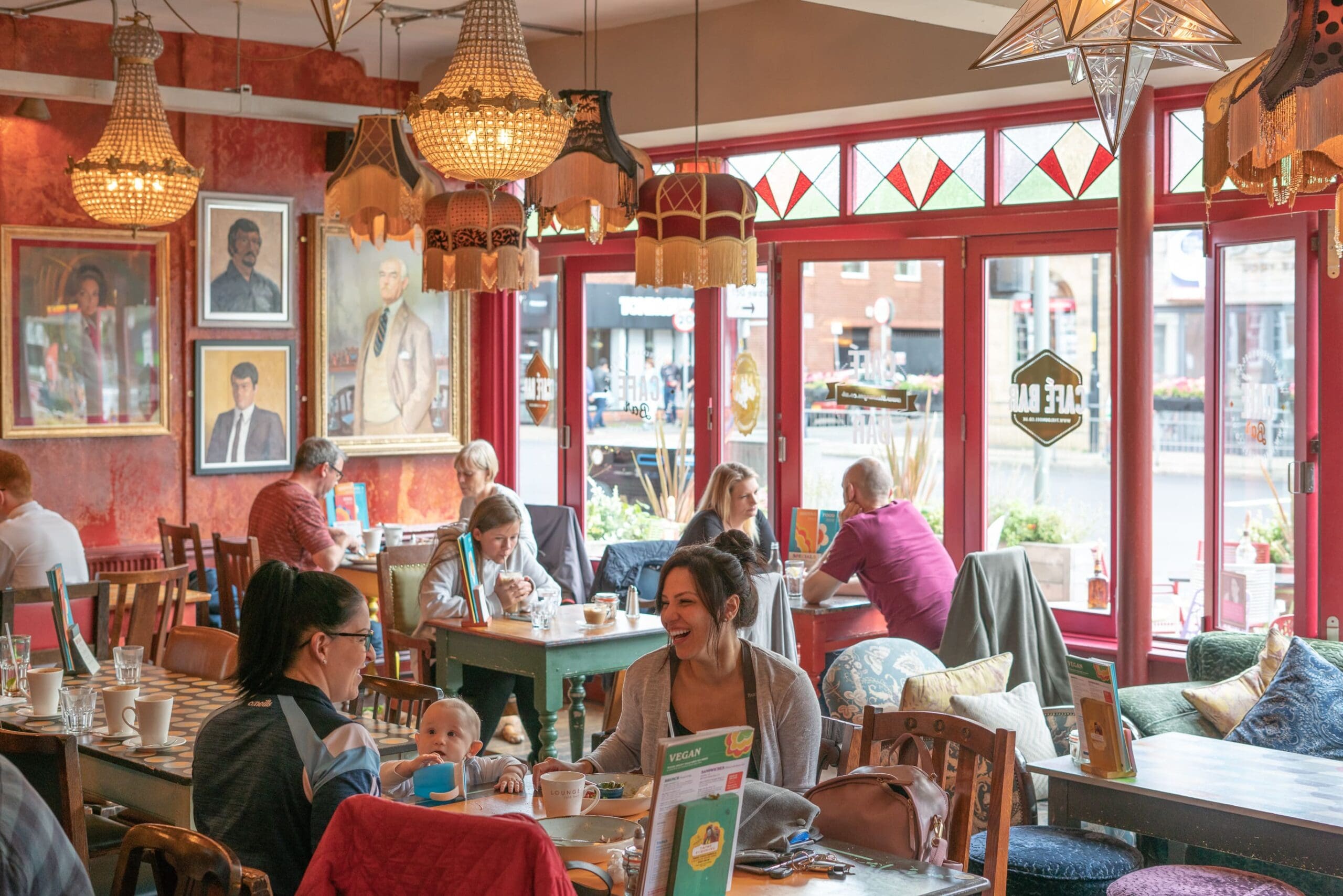 A cozy café with people enjoying food and conversation at colorful tables; large windows let in daylight, and ornate chandeliers hang from the ceiling. Expo-inspired paintings decorate the red walls, creating a warm, inviting atmosphere.