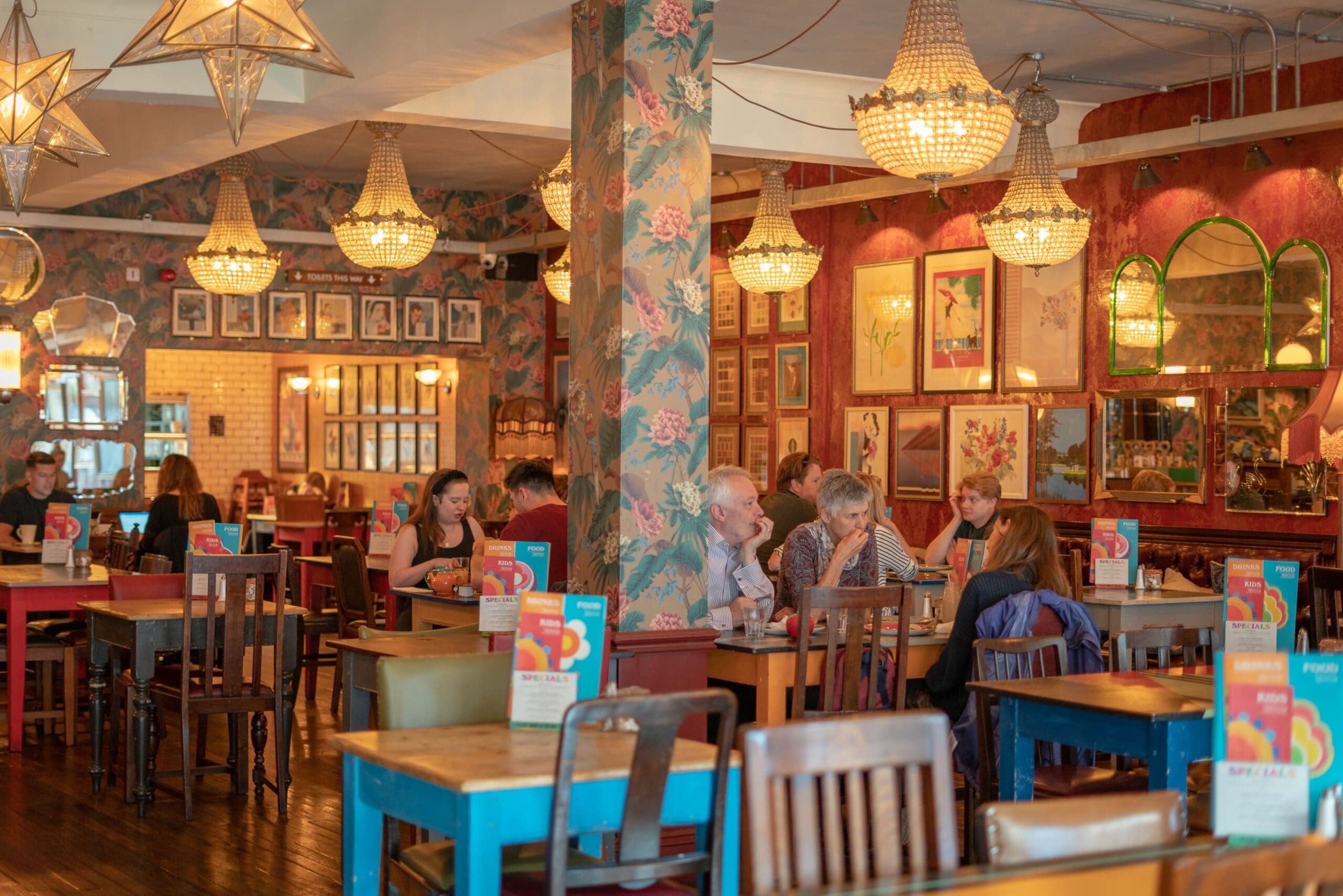 People are dining in a cozy, colorful restaurant with floral wallpaper, vintage chandeliers, and mismatched wooden tables and chairs, creating an atmosphere as lively and welcoming as an Expo. Framed art adorns the walls.